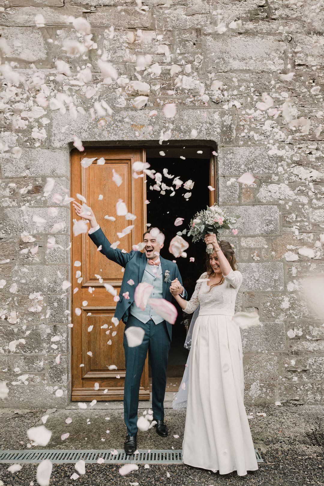 Un couple de mariés joyeux sortant d'une église, lançant des pétales de fleurs blanc et rose, la femme tenant un bouquet et le couple se tenant par la main.
