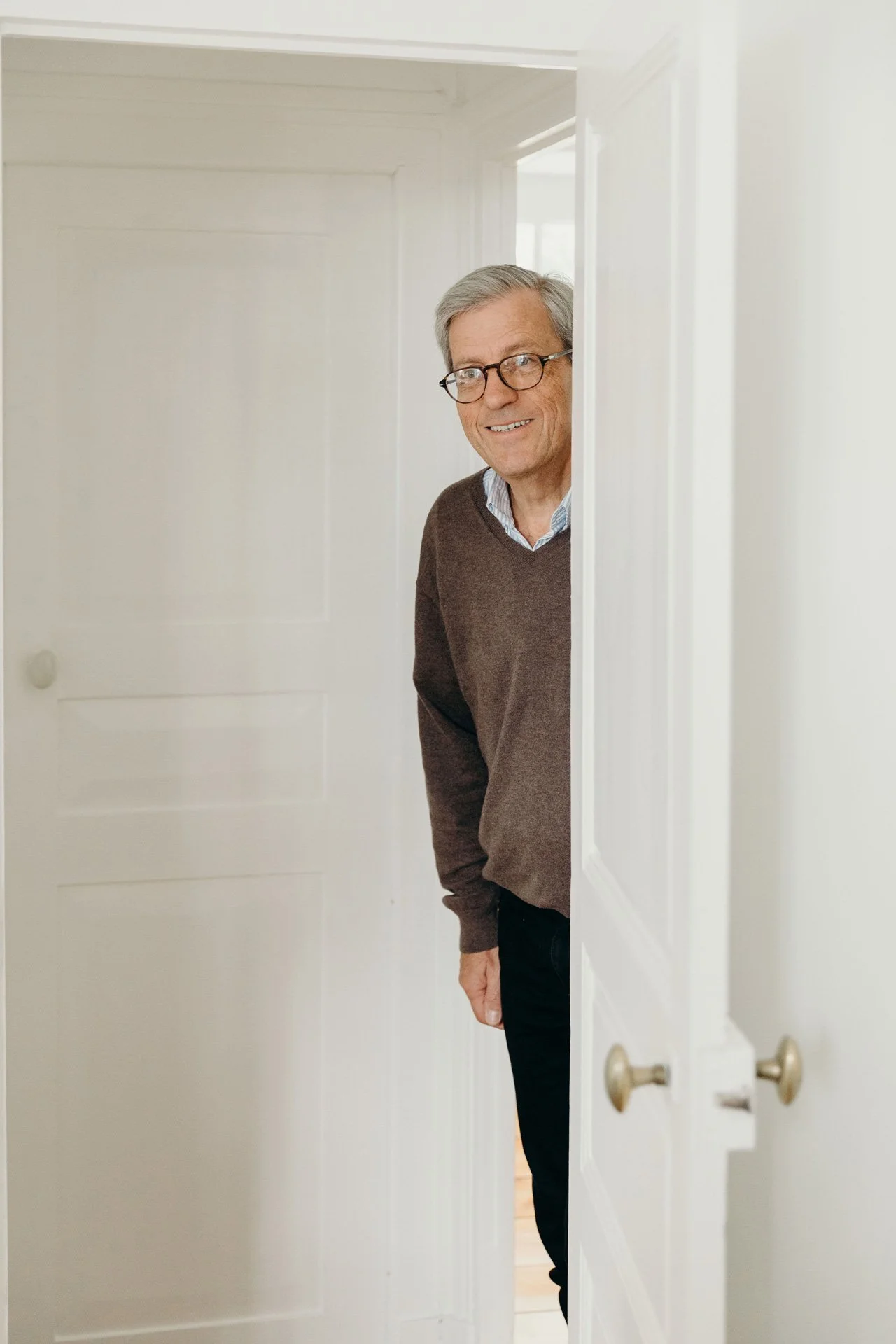 Un homme âgé porte des lunettes, sourit et se penche à travers une porte entrouverte dans une pièce lumineuse.