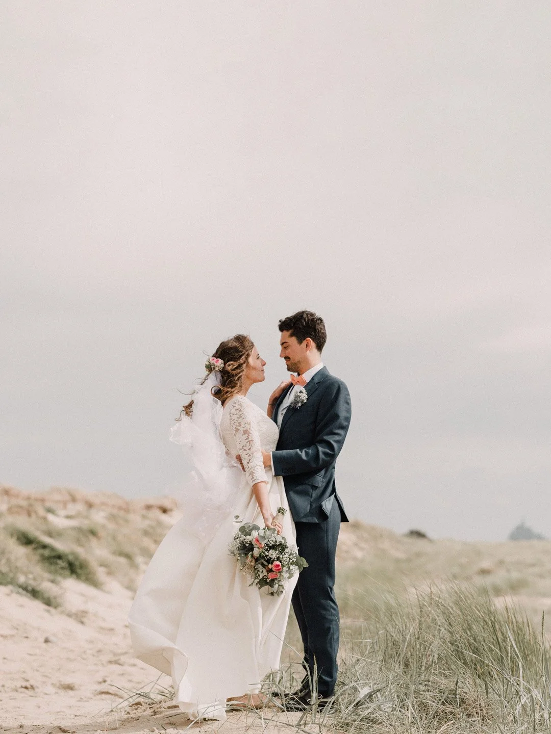 Couple de mariés se tenant par la main et se regardant tendrement dans la baie du Mont-Saint-Michel, paysage naturel et ciel nuageux en arrière-plan.
