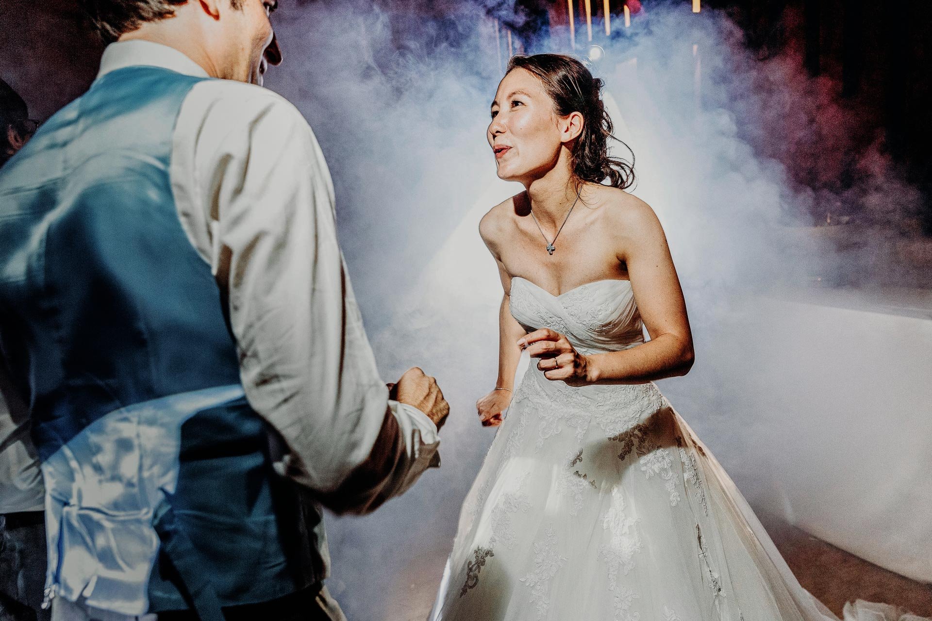 Une femme en robe de mariée et un homme en veston discutent lors d'une réception de mariage, avec un fond de fumée et lumières colorées.