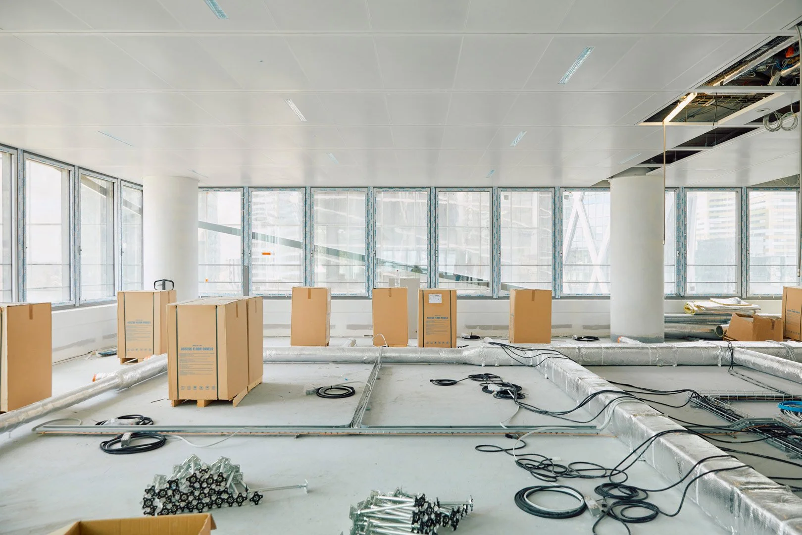 Une pièce en construction avec des fenêtres laissant passer la lumière naturelle, des câbles et des cartons de matériel électrique ou de plomberie, et des équipements en cours d'installation.