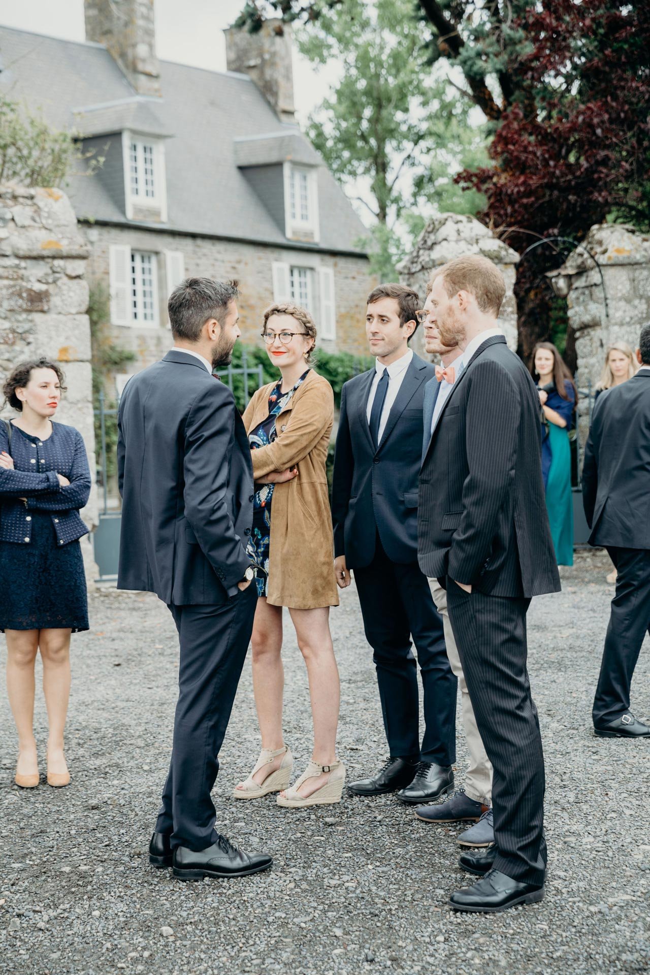 Groupe de personnes en costume lors d'un événement en extérieur devant une maison ancienne en pierre, discutant et souriant.