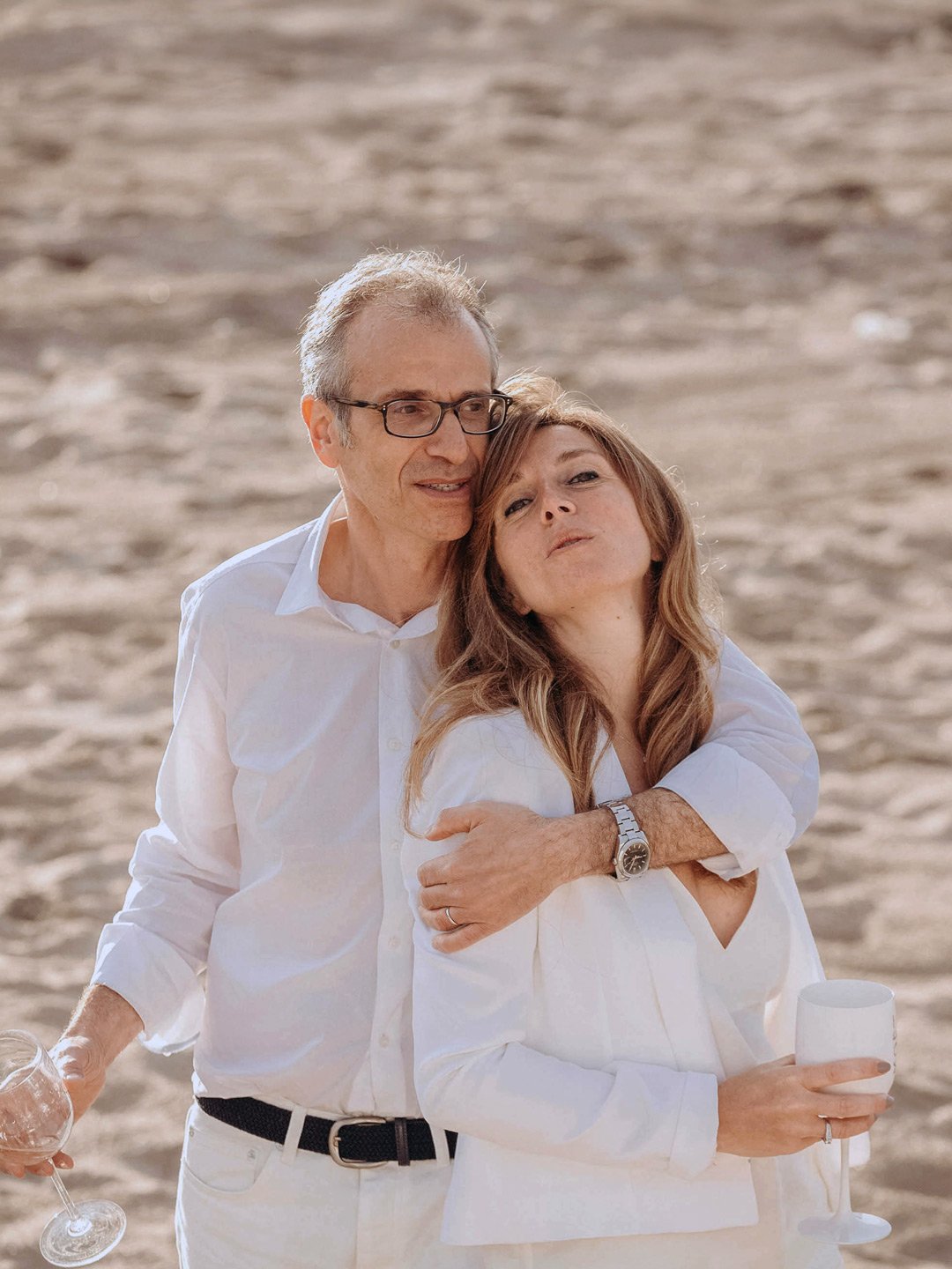 Un homme et une femme en tenue blanche enlassés sur la plage de Deauville, la femme tient un verre, l'homme un verre à vin, arrière-plan de sable.