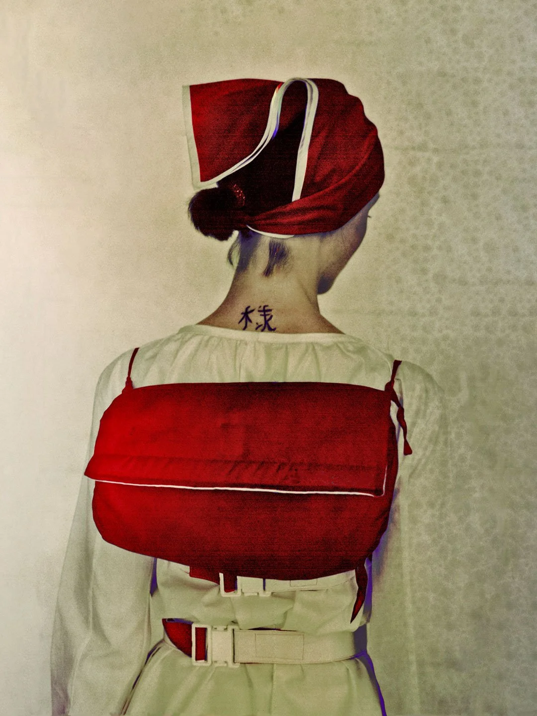 Personne vue de dos portant un bonnet rouge avec un toupie noire, un sac rouge, un haut blanc, et une tatouage chinoise au dos