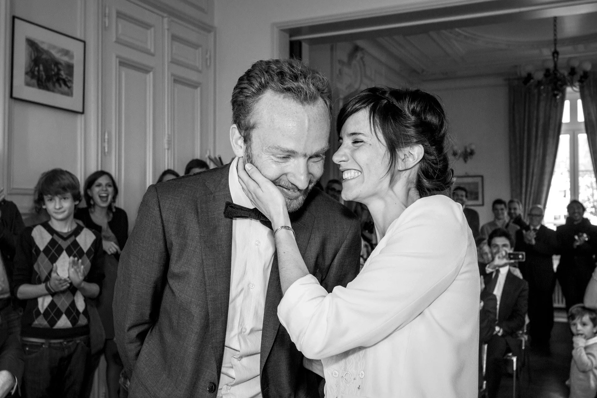 Un couple souriant lors d'une cérémonie civile avec des invités en arrière-plan, en noir et blanc.