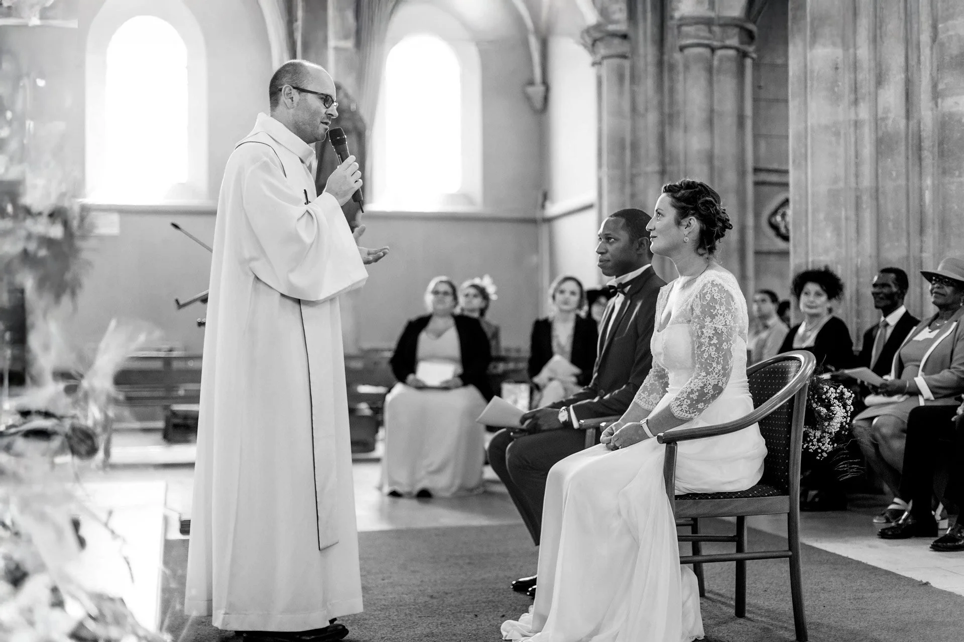 Un prêtre officiant un mariage dans une église avec un couple assis lors de la cérémonie et des invités en arrière-plan, en noir et blanc.