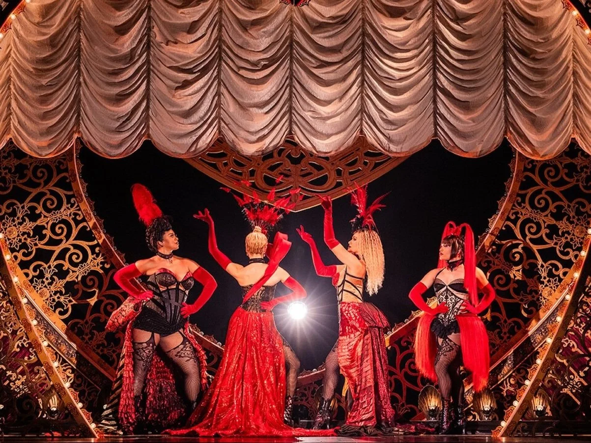 Four women dressed in showgirl costumes on stage with ornate gold and red decorations, large curtains, and dramatic lighting.