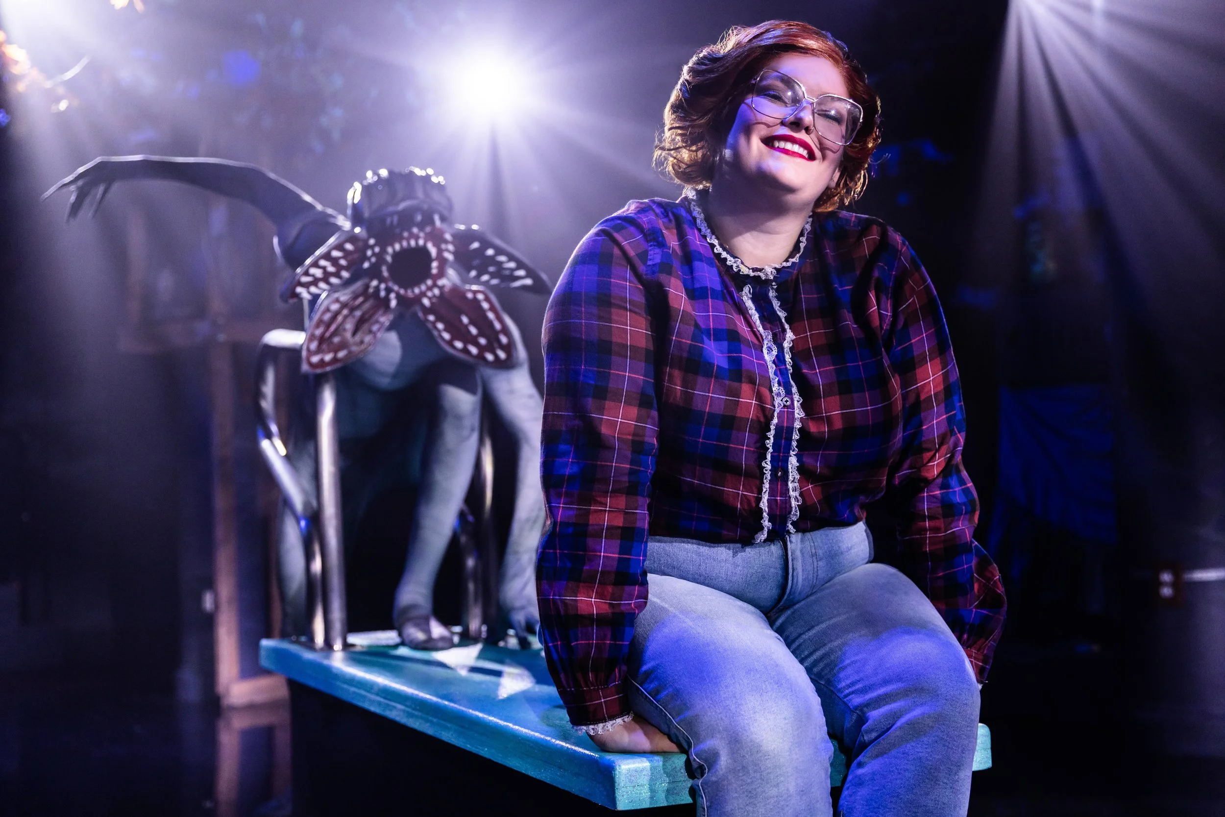 A woman with short brown hair and glasses sits on a bright blue platform, smiling. She wears a red and blue plaid shirt and light blue jeans. Behind her, a performer in a costume with a large flower mask and bat-like wings crouches on the platform, under bright stage lights.