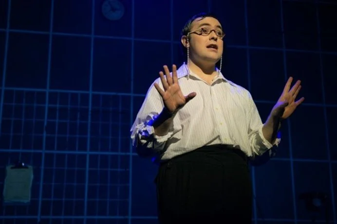 A woman with glasses, wearing a white striped shirt, gesturing with her hands while speaking or performing on stage against a blue tiled background.