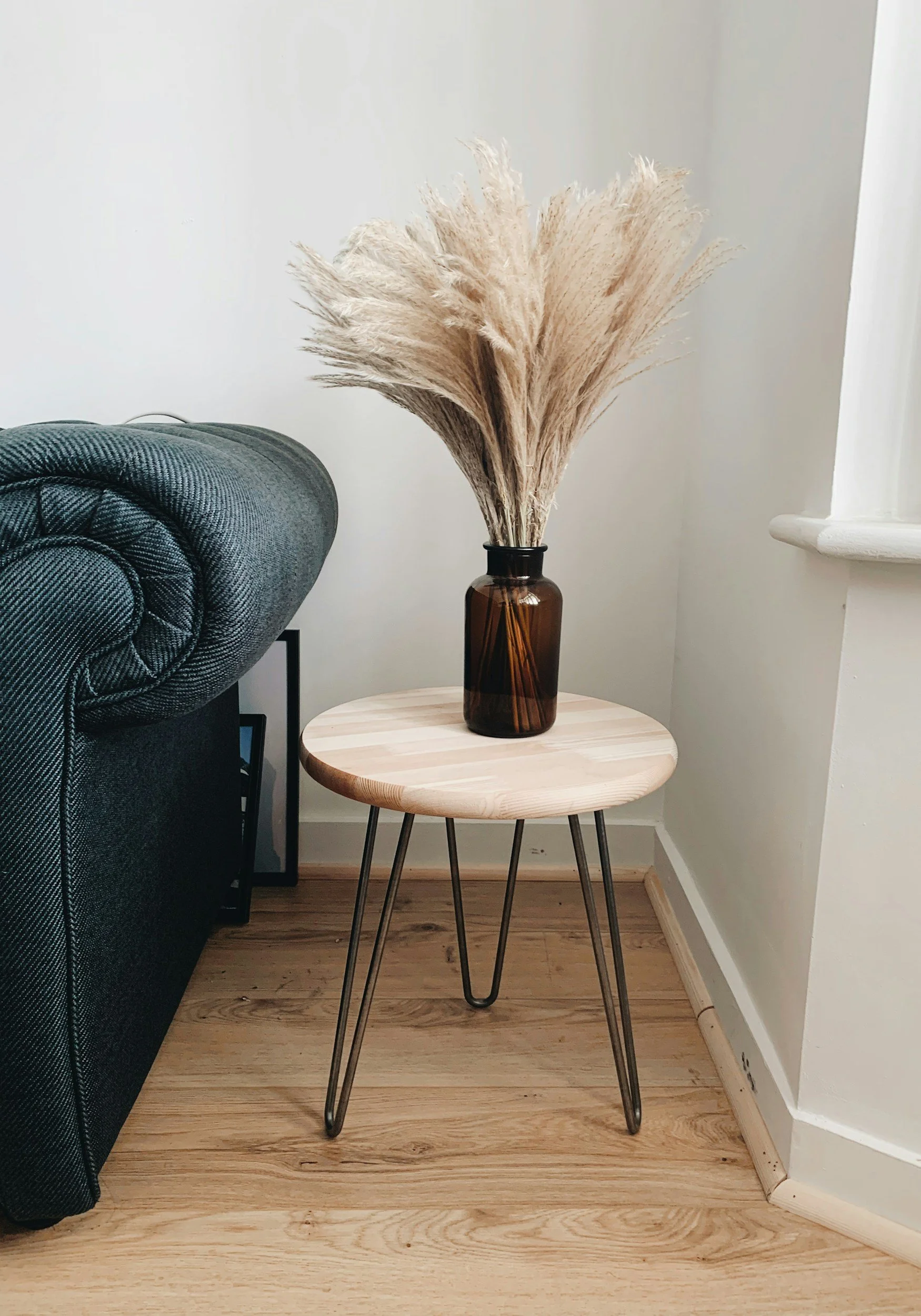 A round wooden side table with black metal hairpin legs, holding a brown glass vase filled with dried pampas grass, next to a green upholstered sofa in a corner of a room with light-colored walls and wood flooring.