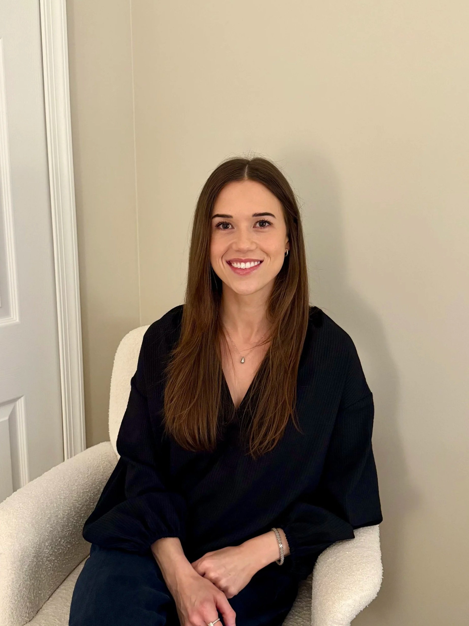 A woman with long brown hair, smiling, wearing a black blouse, sitting on a white armchair in a room with beige walls.