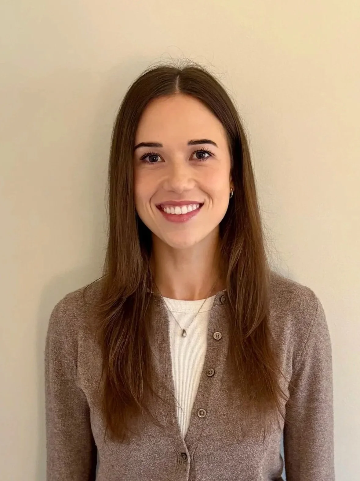 A woman with long brown hair, smiling, wearing a beige cardigan over a white top and a necklace with a pendant, standing against a plain light-colored background.