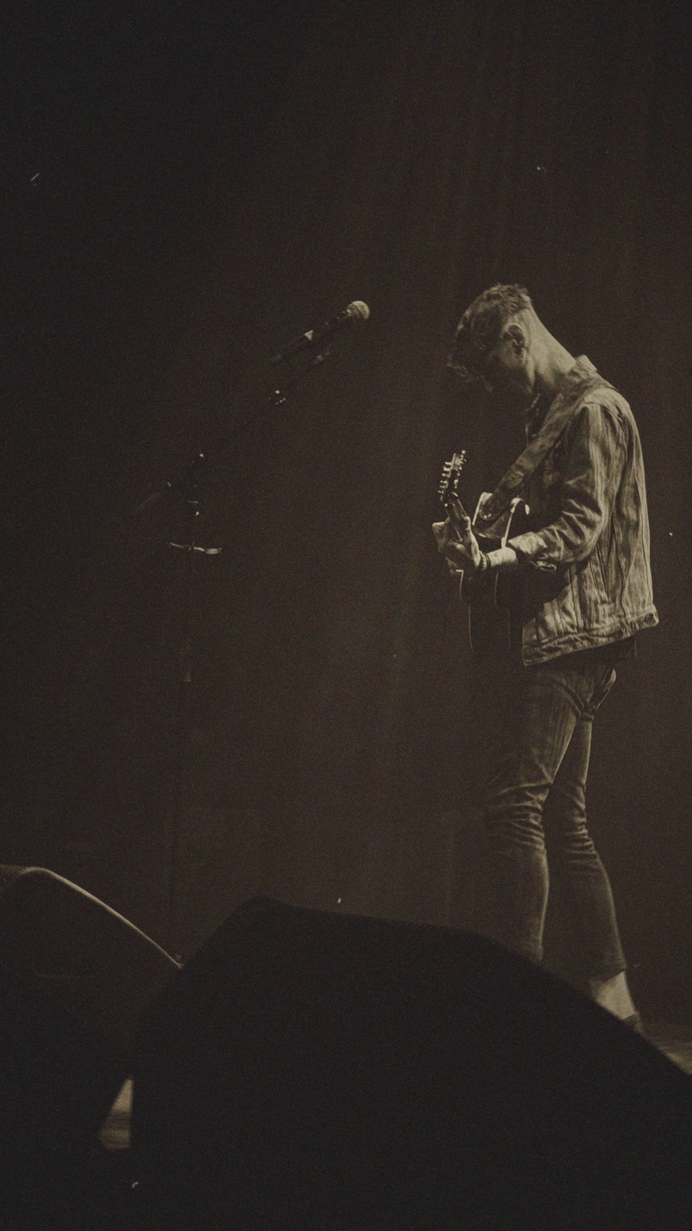 A musician playing an electric guitar on stage, illuminated by dim lighting, wearing glasses and a jacket.