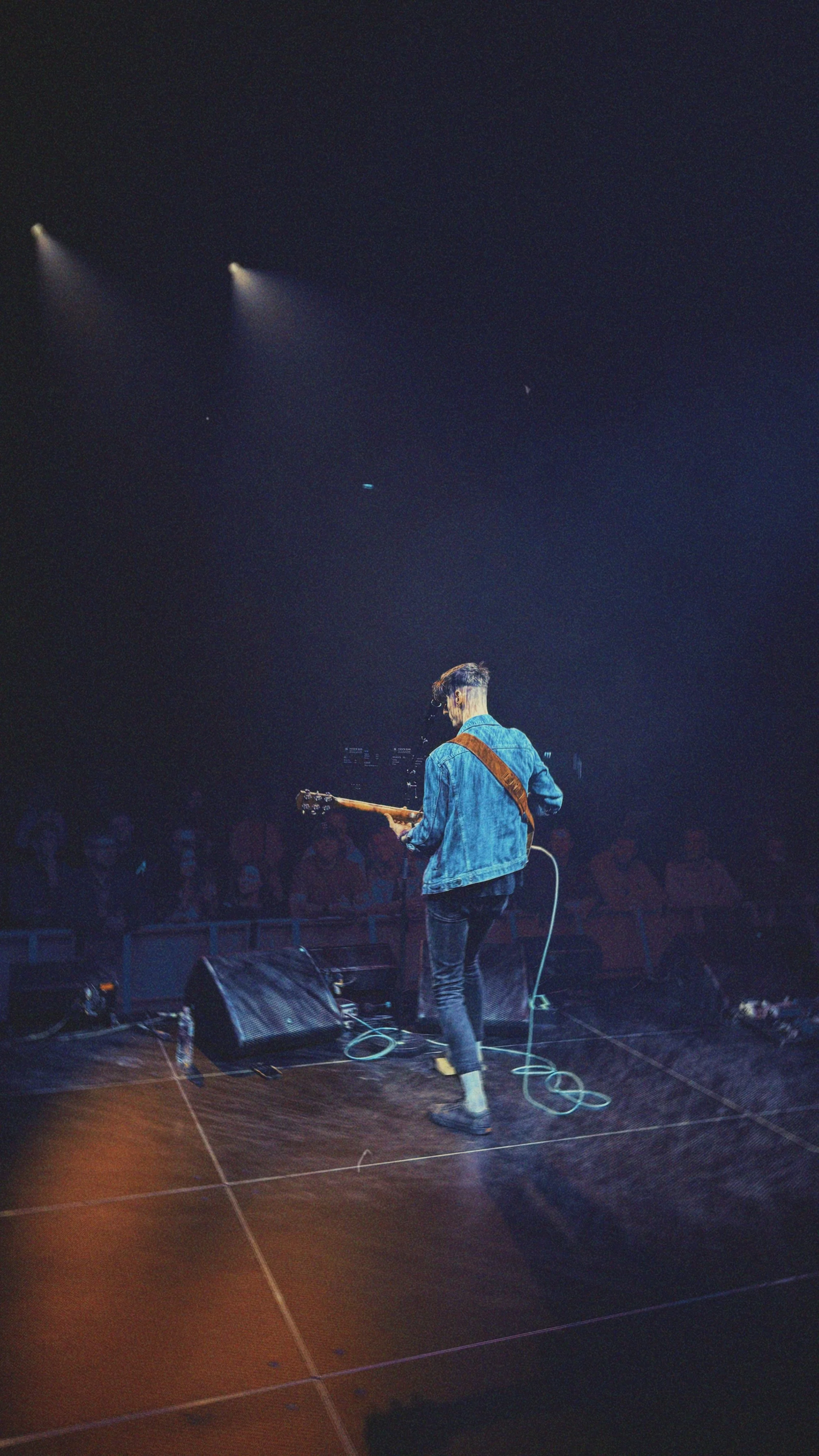 A person with short hair wearing a blue denim jacket and black pants, playing an electric guitar on stage in front of an audience, with stage lights overhead.