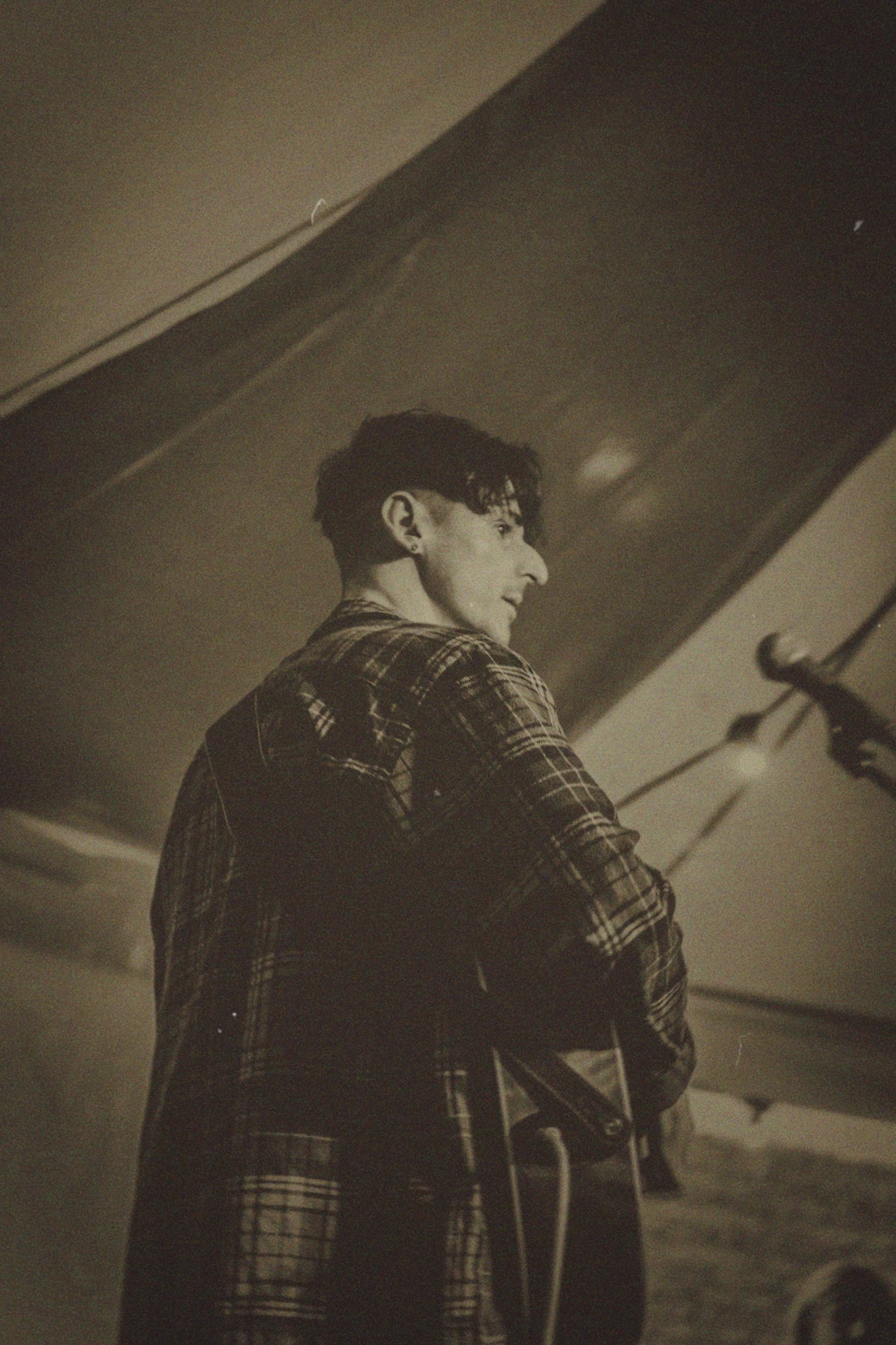 A person with short dark hair and earrings, wearing a plaid shirt, standing near an airplane and looking to the side.