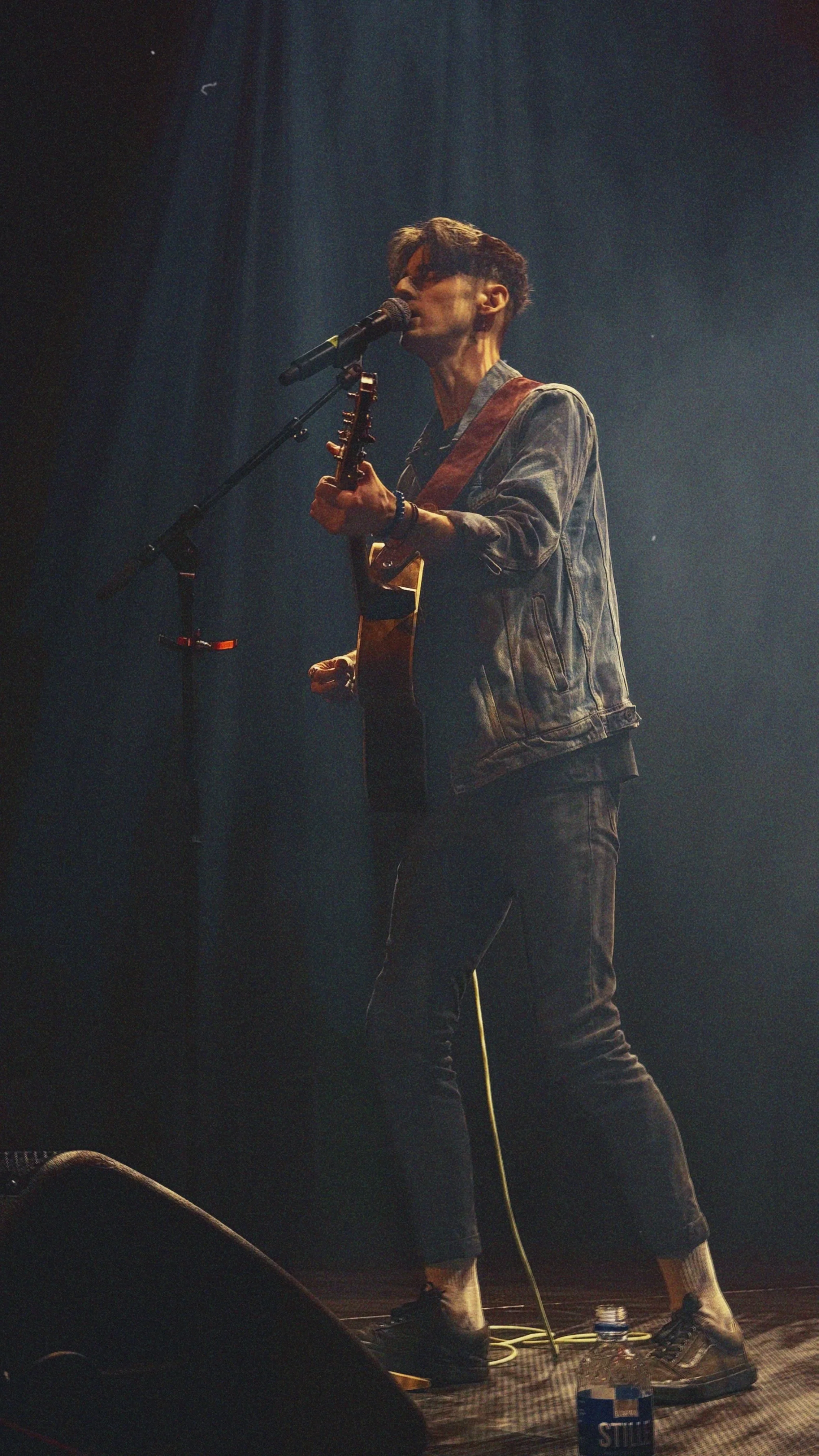 A male musician with short brown hair performing on stage, singing into a microphone and playing an acoustic guitar, wearing a denim jacket, black pants, and sneakers, with stage lights shining down and a water bottle on the floor.