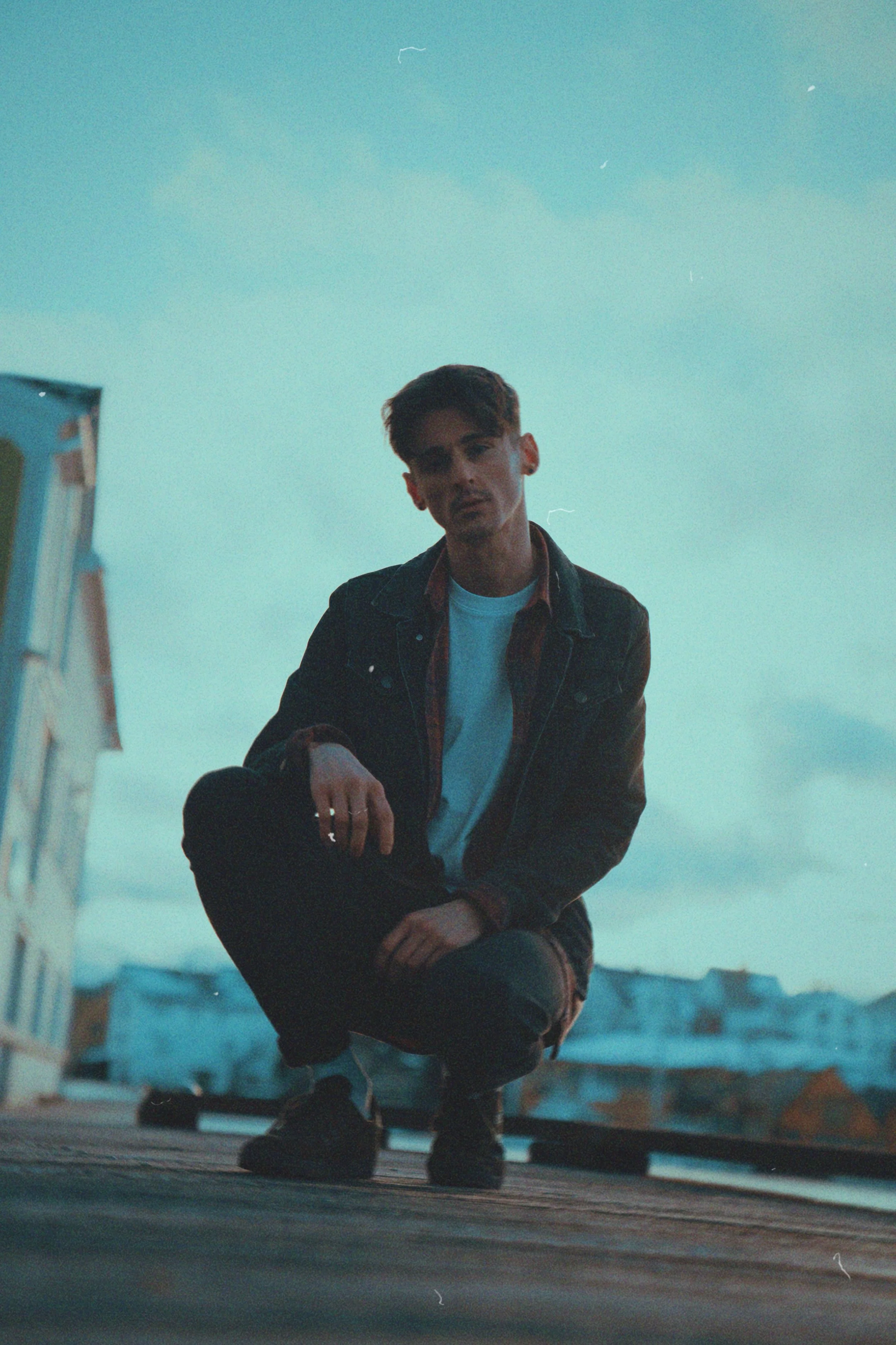 A young man with dark hair and styled hair is squatting on a rooftop, looking at the camera, wearing a denim jacket over a checked shirt and black shoes against a teal sky background at dusk.