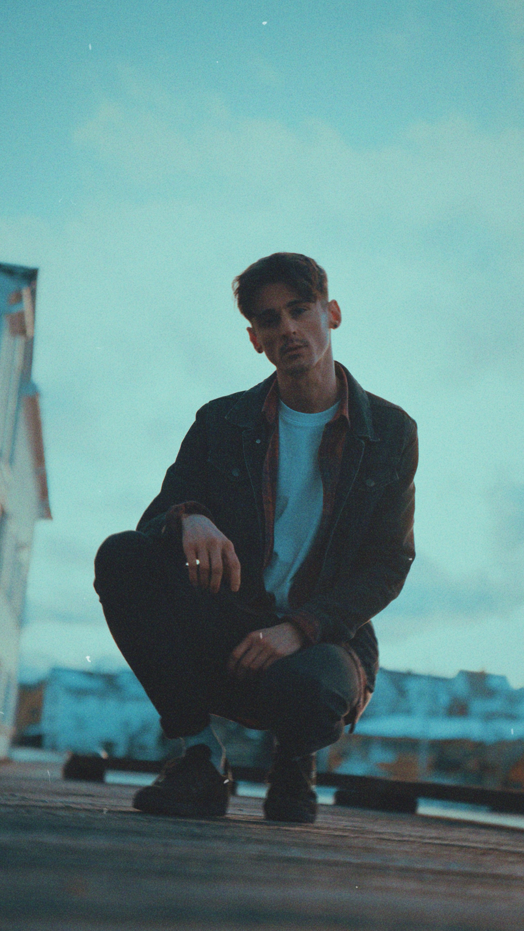 A young man with styled hair and dark clothing crouches on a rooftop at dusk, with a city skyline and overcast sky in the background.