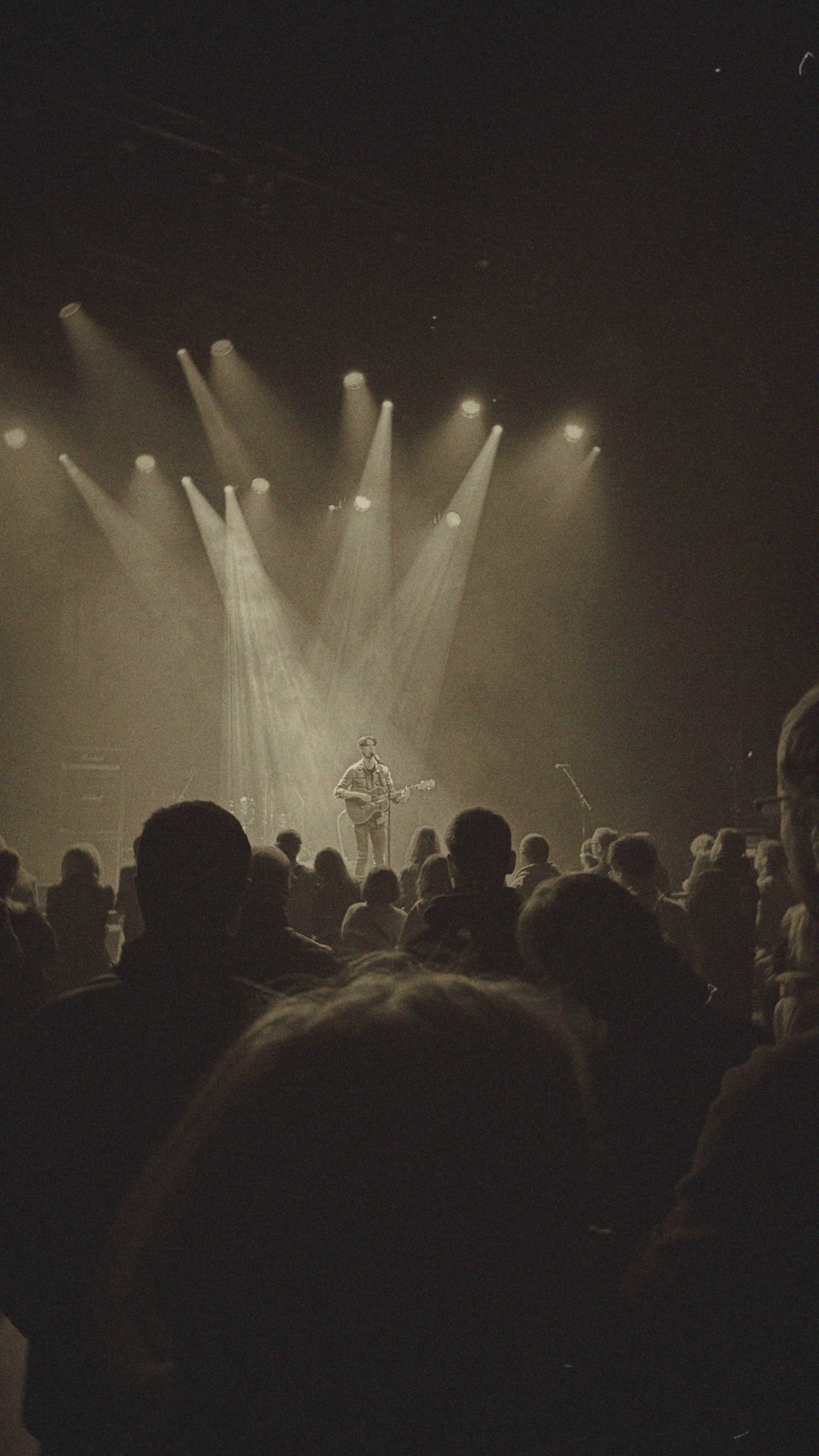 A musician playing guitar on stage under bright stage lights with an audience watching.
