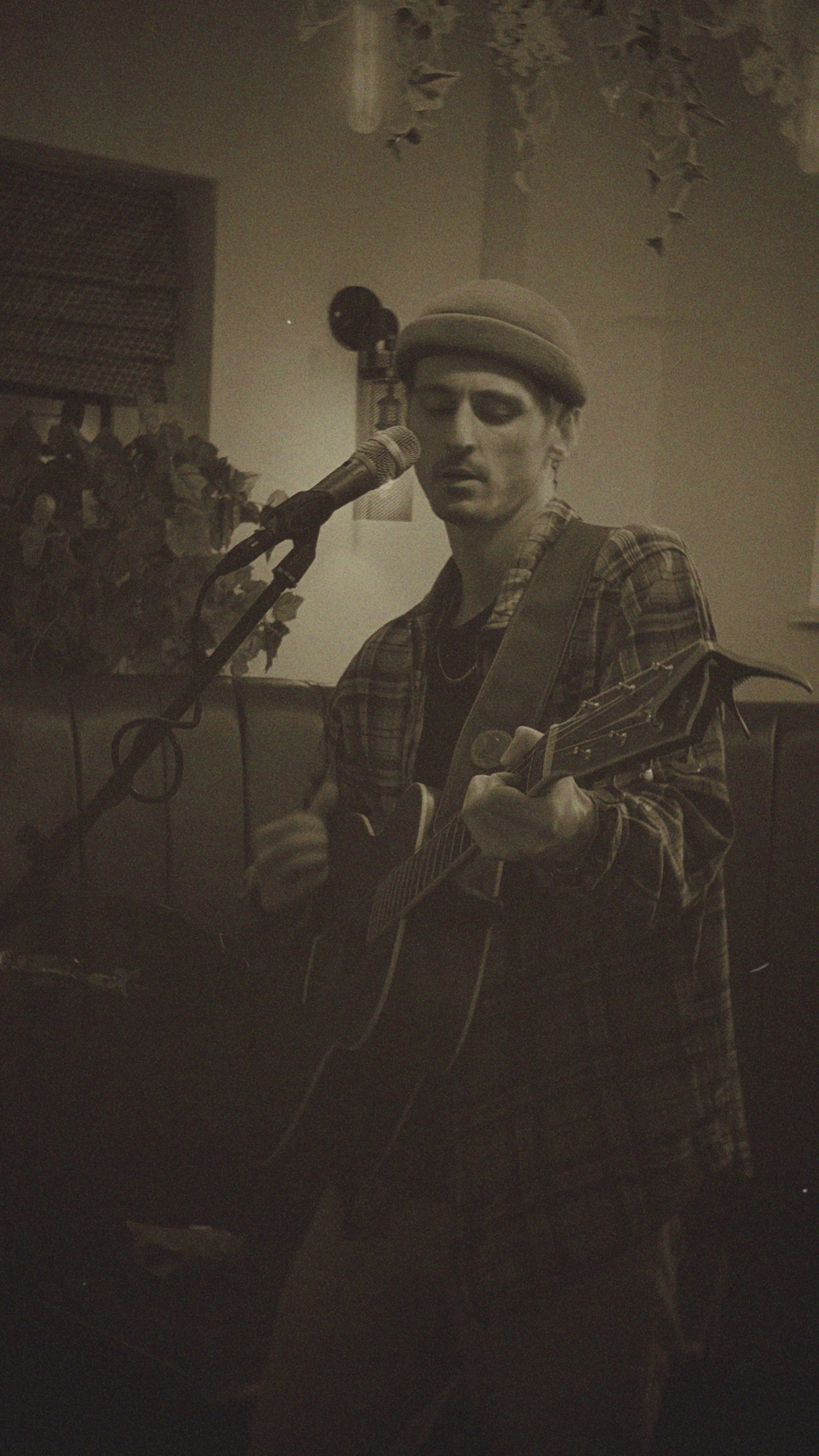 A person wearing a plaid shirt and a beret is singing into a microphone while playing an acoustic guitar, in a dimly lit room with leaves hanging from the ceiling.