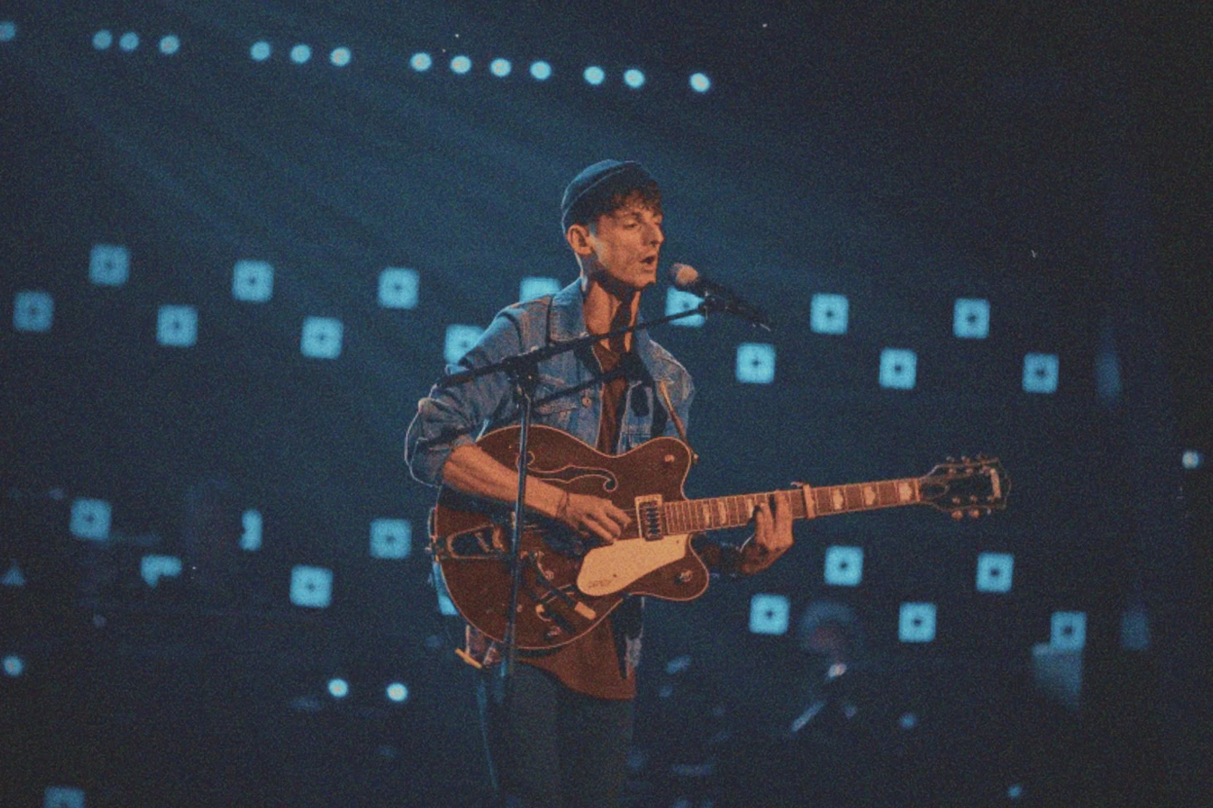 Musician playing guitar and singing into a microphone on stage with blue and white light backdrop.