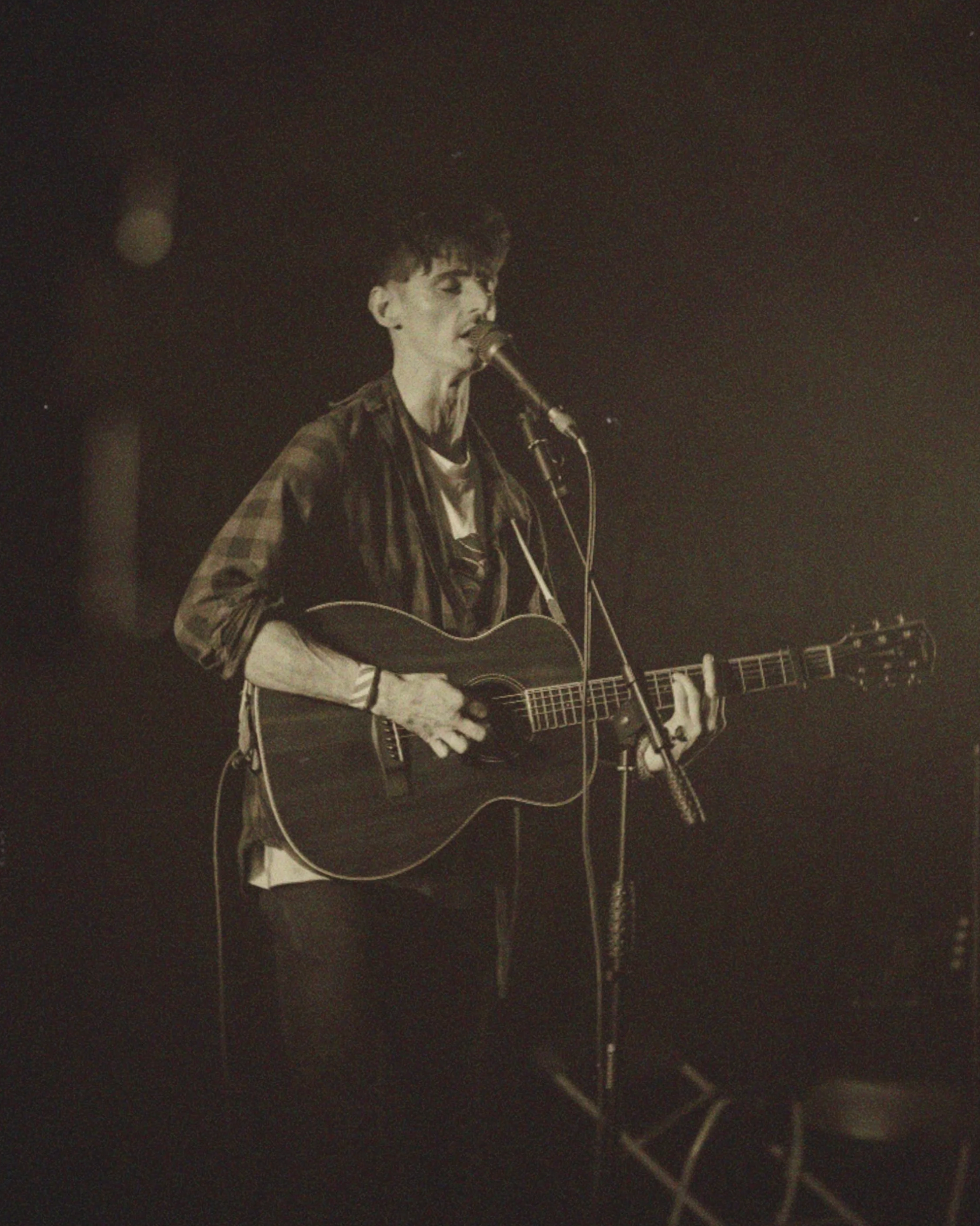 A man with short curly hair playing an acoustic guitar and singing into a microphone on a dark stage.