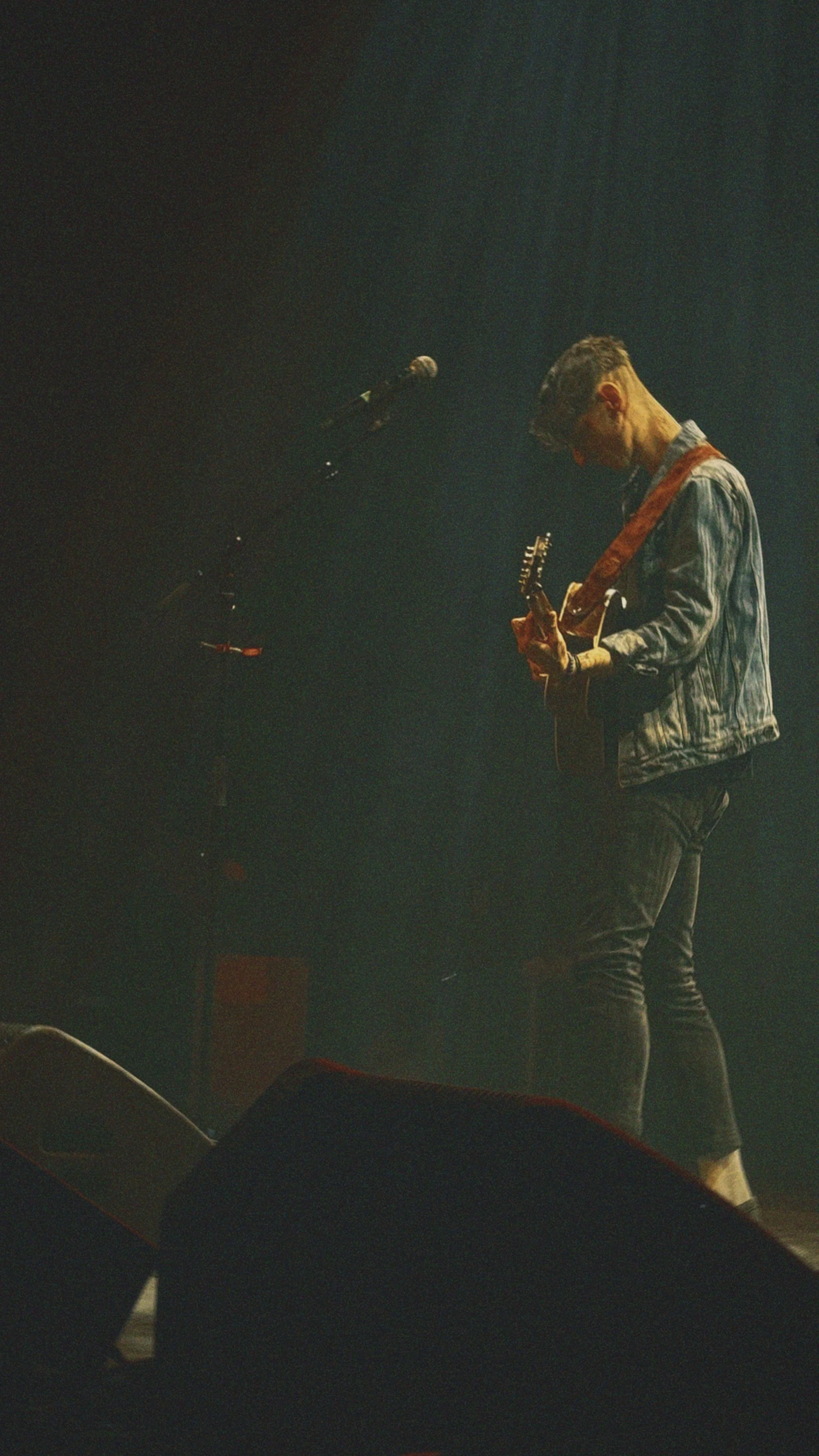 A male musician playing guitar on stage during a live performance, with a microphone stand nearby and stage lighting creating a smoky atmosphere.
