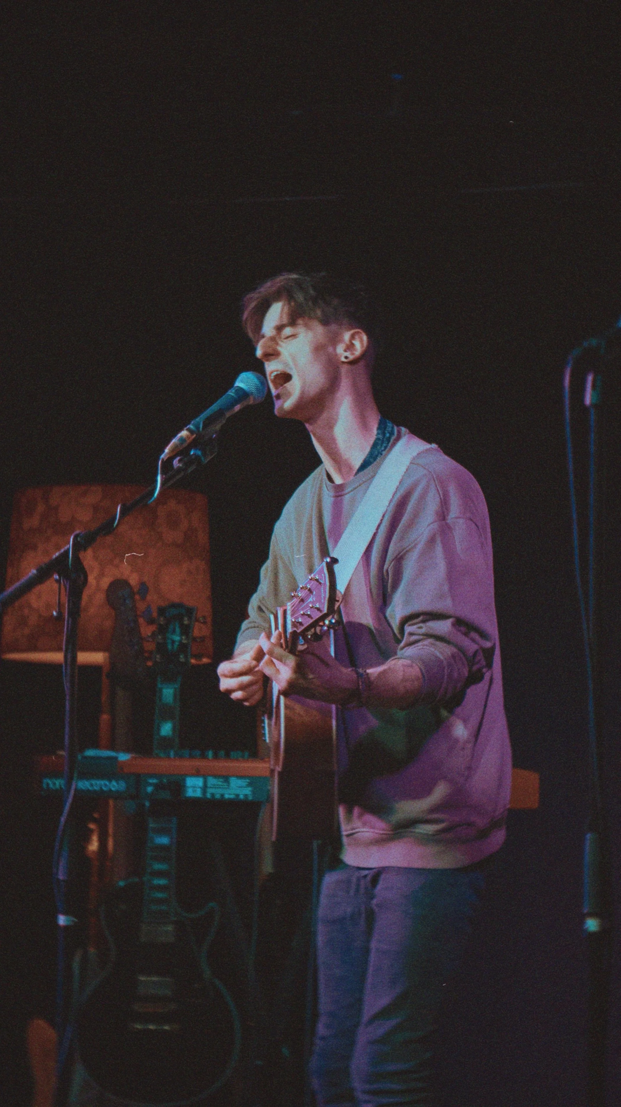 A young man performing on stage plays guitar and sings into a microphone with closed eyes, wearing a beige sweatshirt, in a dimly lit venue with sound equipment and a patterned backdrop.