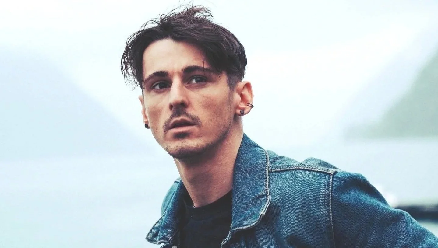 A young man with dark hair, earrings, and a piercings, wearing a denim jacket, looking into the camera with a serious expression, outdoors on a cloudy day.