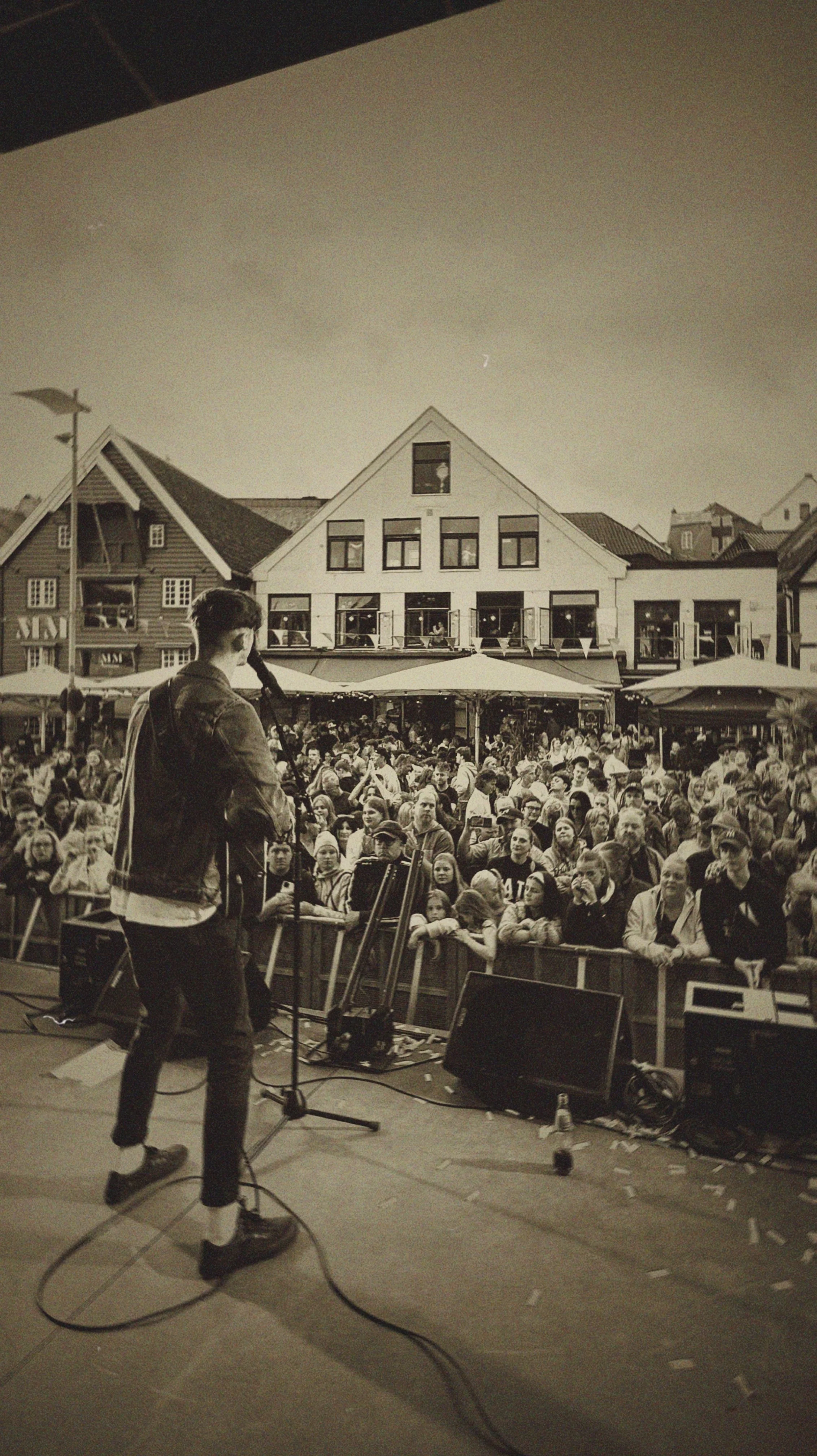 Male musician performing on stage with a microphone in hand, facing a large crowd at an outdoor event, with buildings in the background.