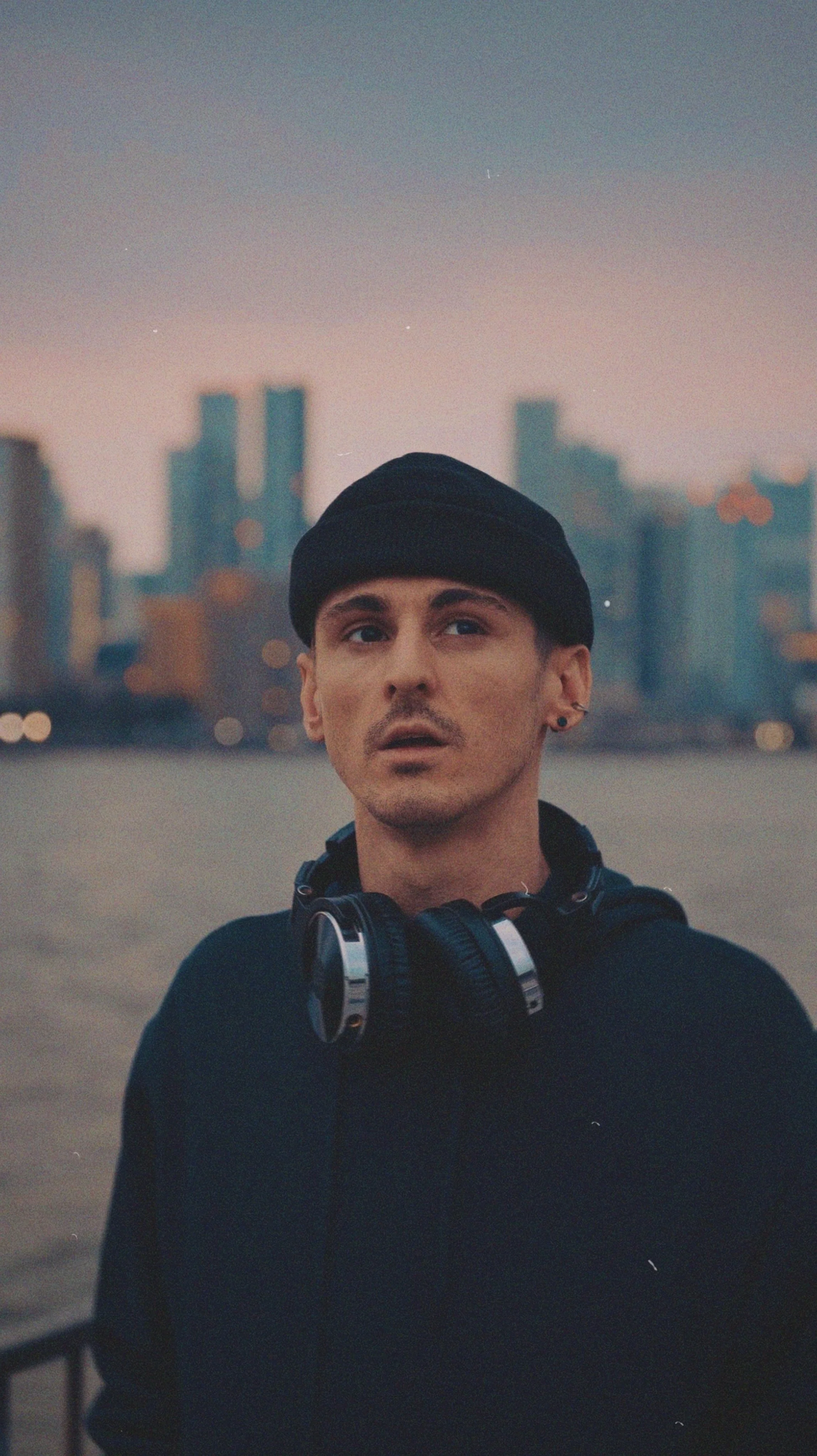 A young man with short dark hair, wearing a black beanie, black hoodie, and headphones around his neck, standing outdoors with a city skyline in the background during twilight.