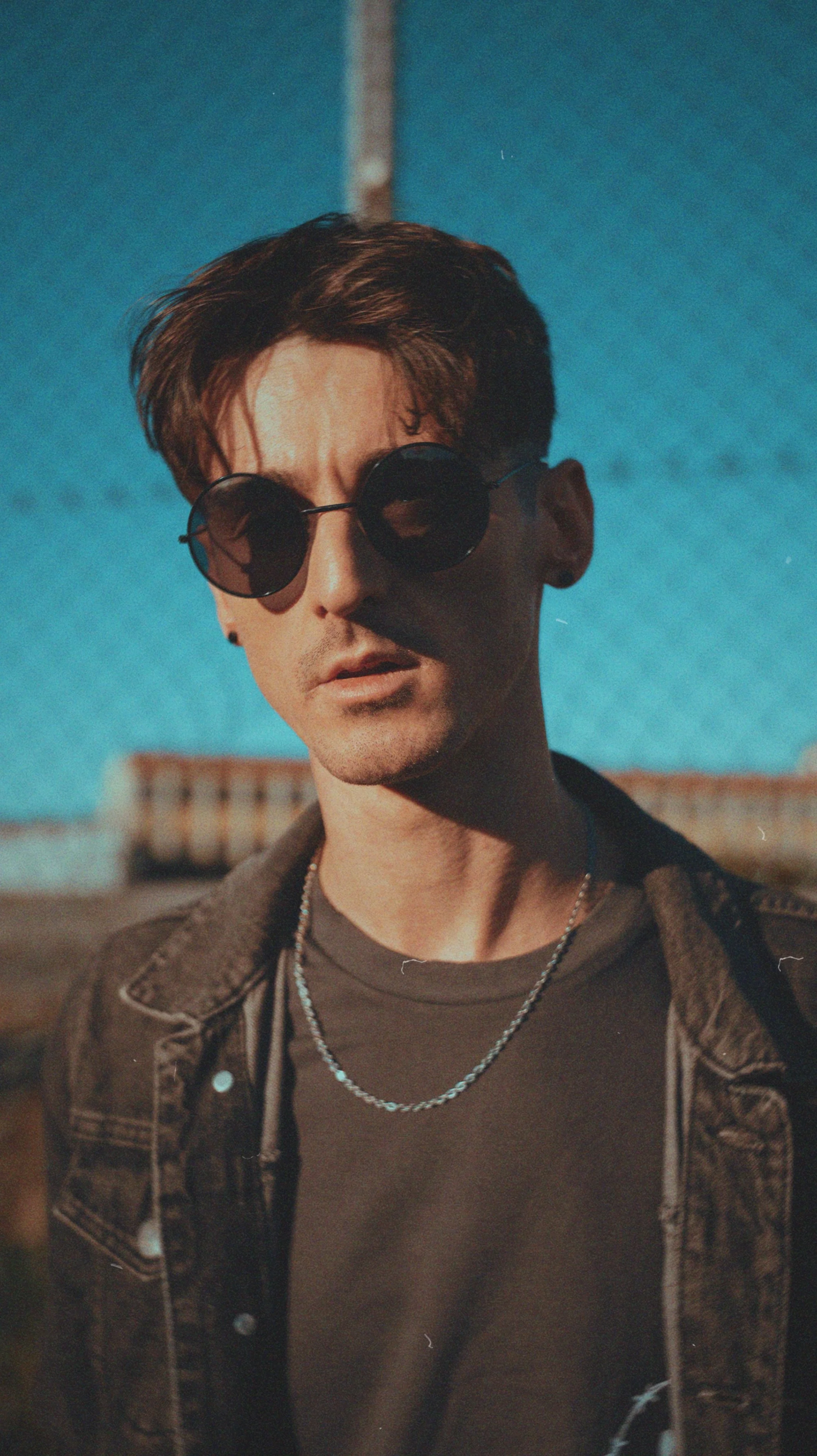 A young man with brown hair wearing round sunglasses, a dark brown shirt, a chain necklace, and a brown jacket standing outdoors with a blue sky and a building in the background.