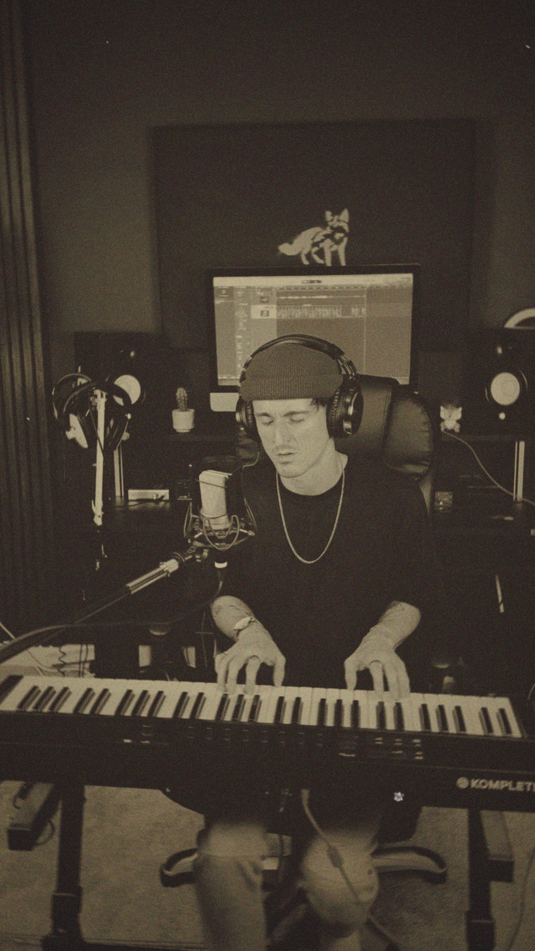 A person in a recording studio, wearing a beanie and headphones, playing a keyboard while singing into a microphone. There are studio monitors, a computer screen, and a wolf decal on the wall behind them.