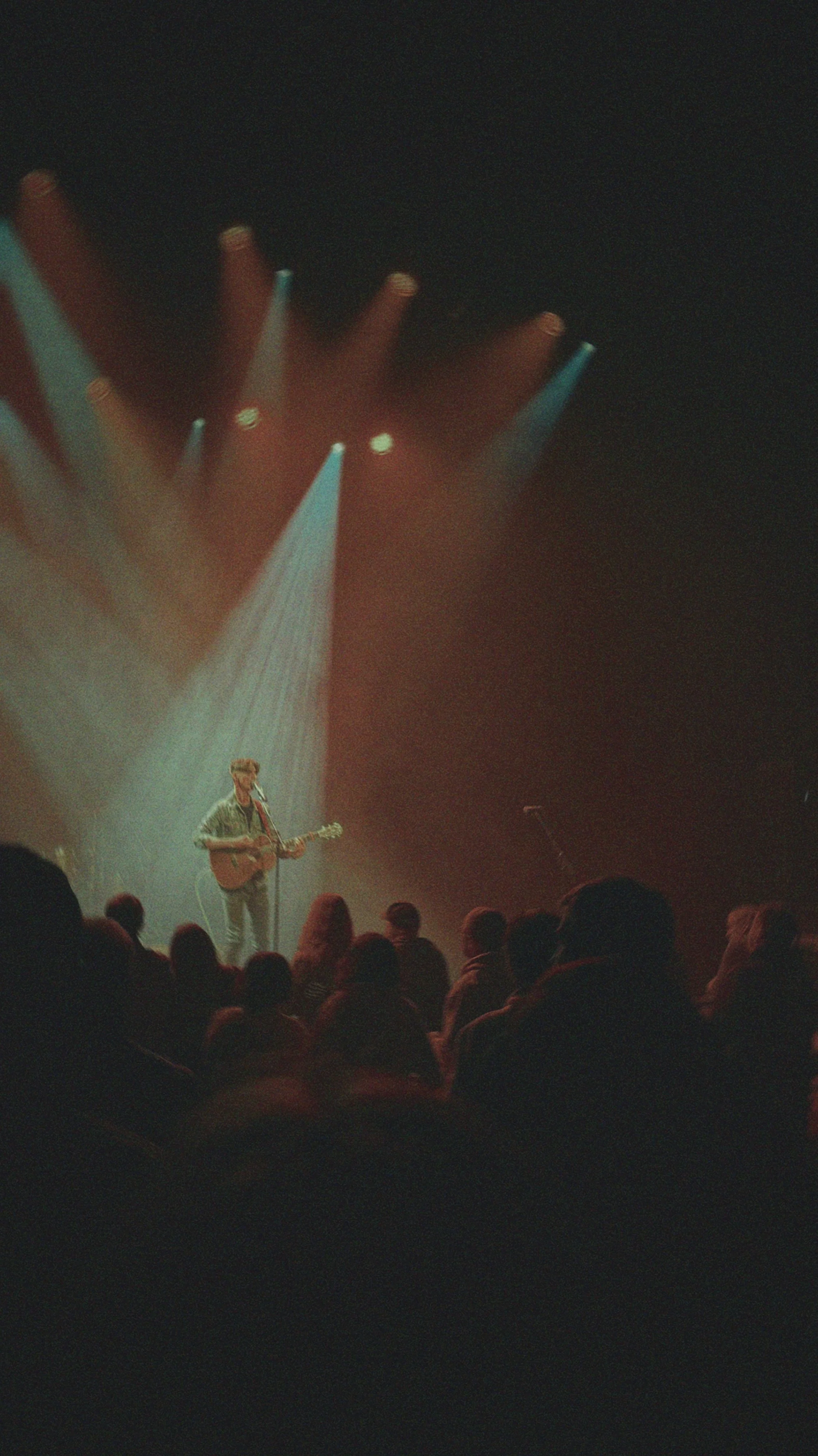 A musician with a guitar performing on a stage during a concert with colorful stage lighting and a crowd in the foreground.