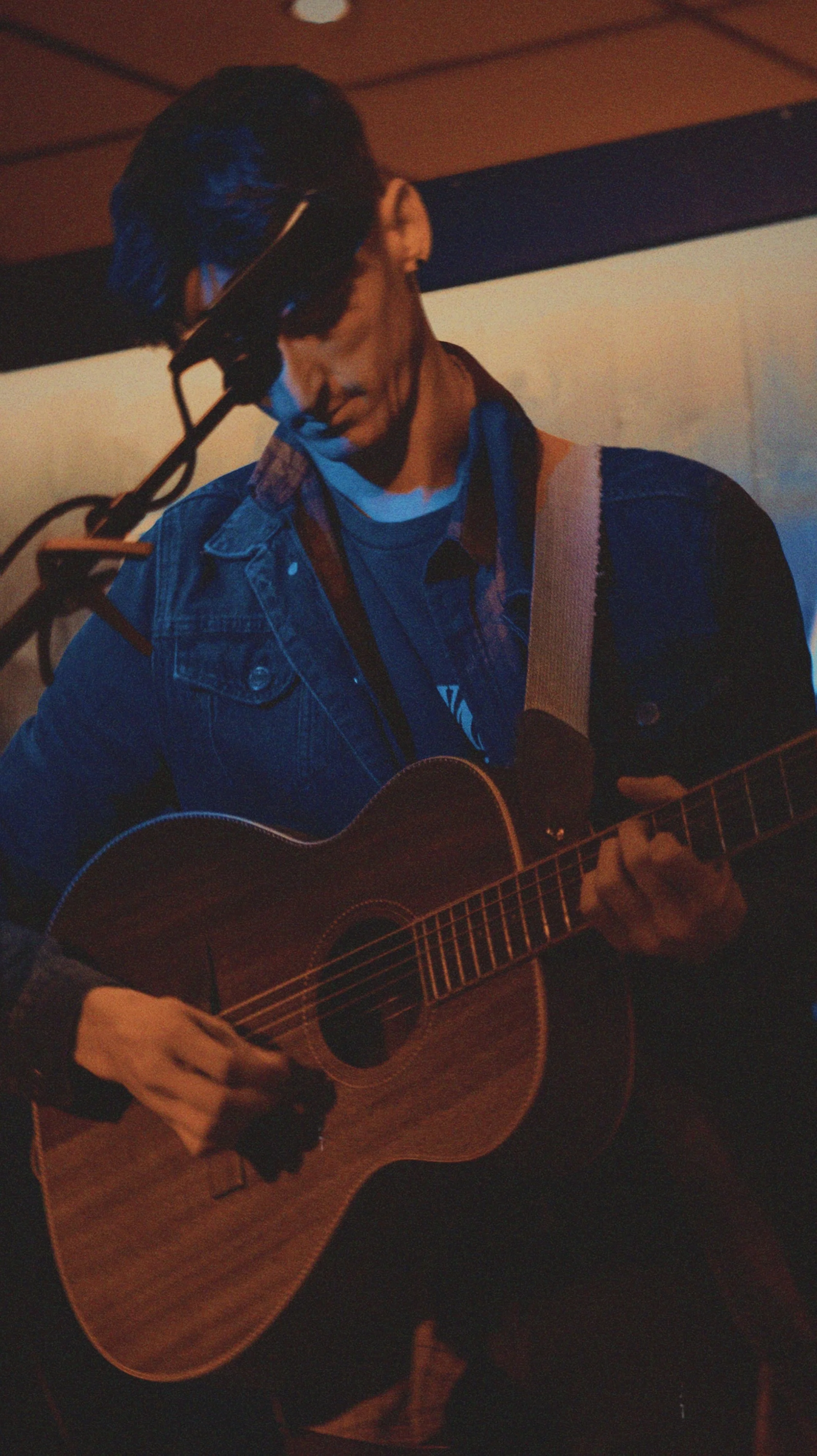 A person playing an acoustic guitar, wearing a denim jacket and a cap, in a dimly lit indoor setting.