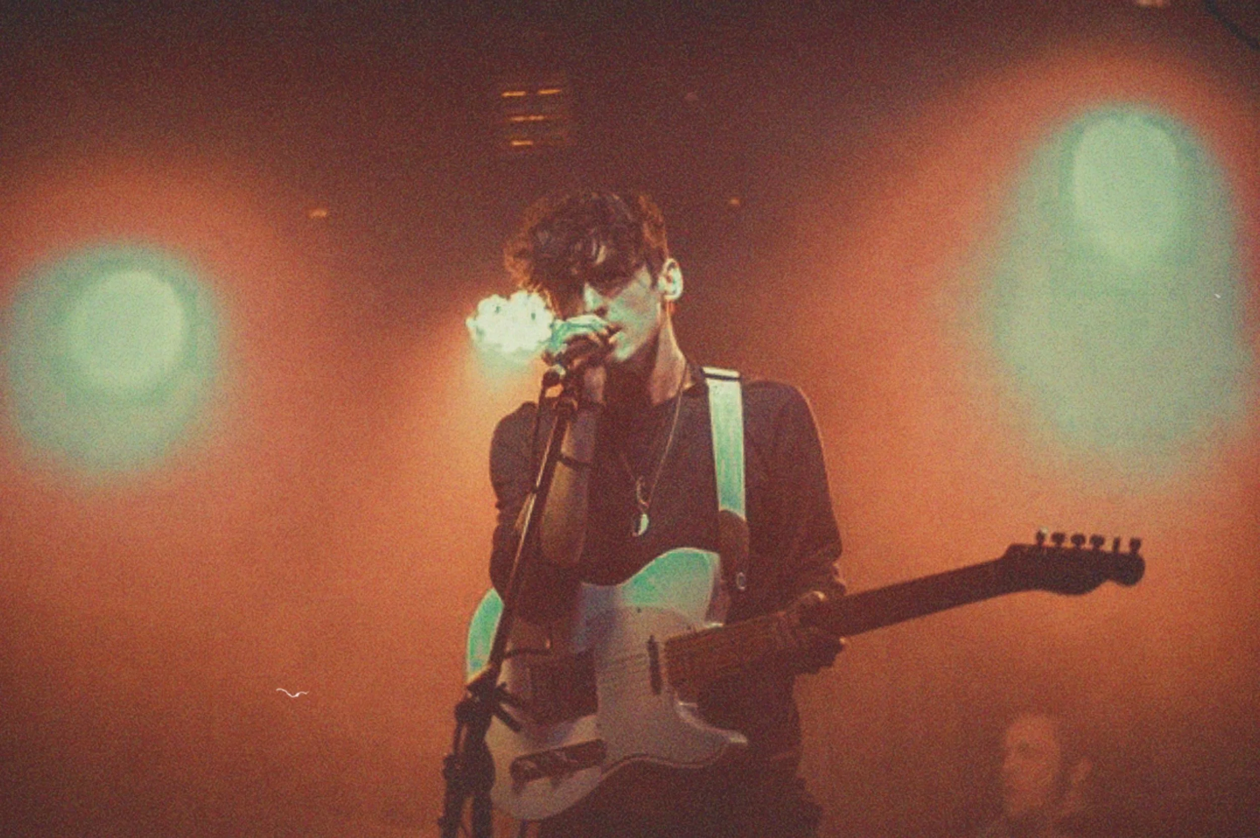 A person with curly hair, wearing glasses and a black shirt, playing an electric guitar on stage with orange lighting, holding a microphone close to their mouth.