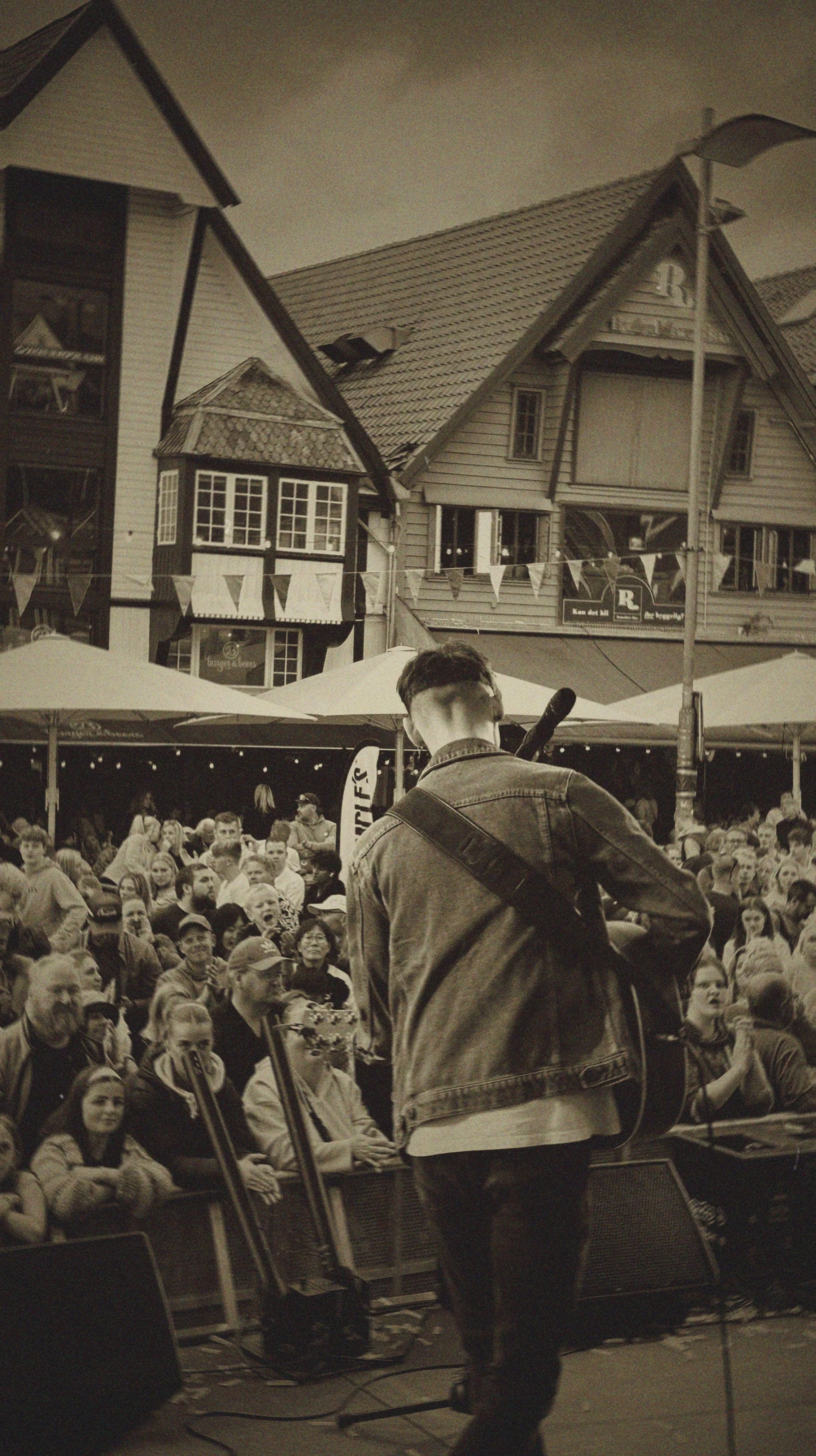 A performer with a guitar on stage at an outdoor concert with a crowd of spectators in front, in front of scenic village-style buildings with pitched roofs.