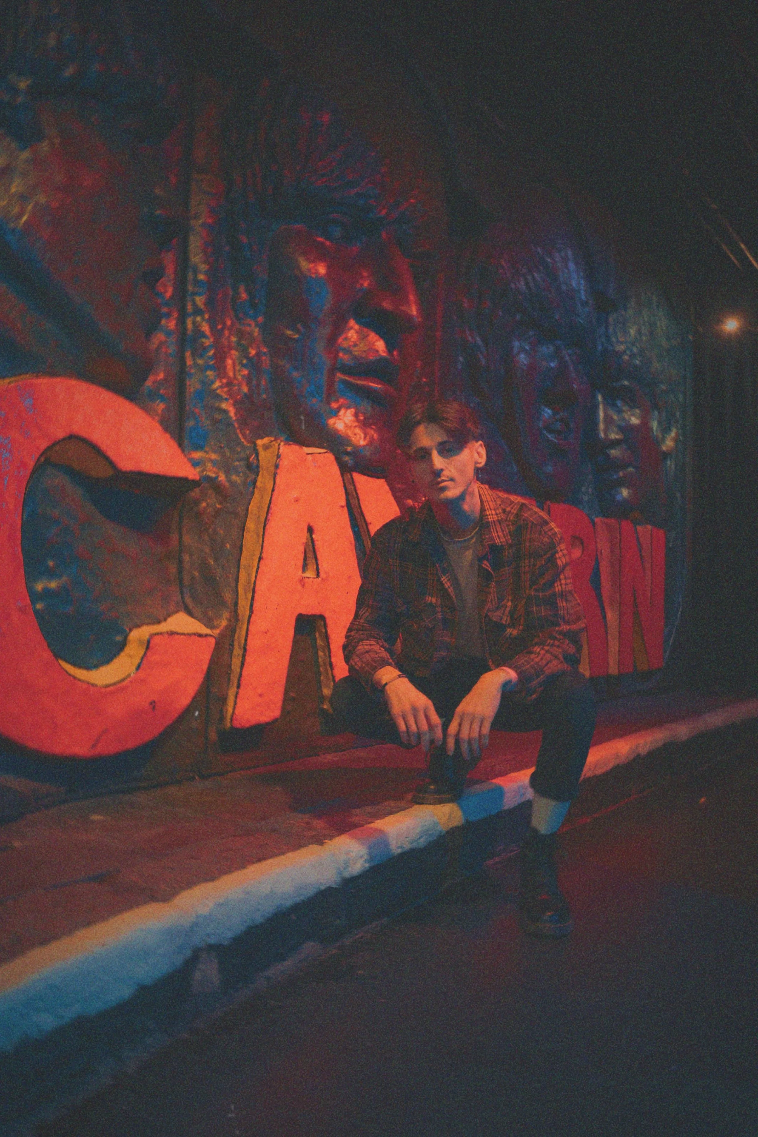 A young man with red hair, wearing a red plaid shirt, sitting on a curb in front of a mural of large, colorful faces at night.
