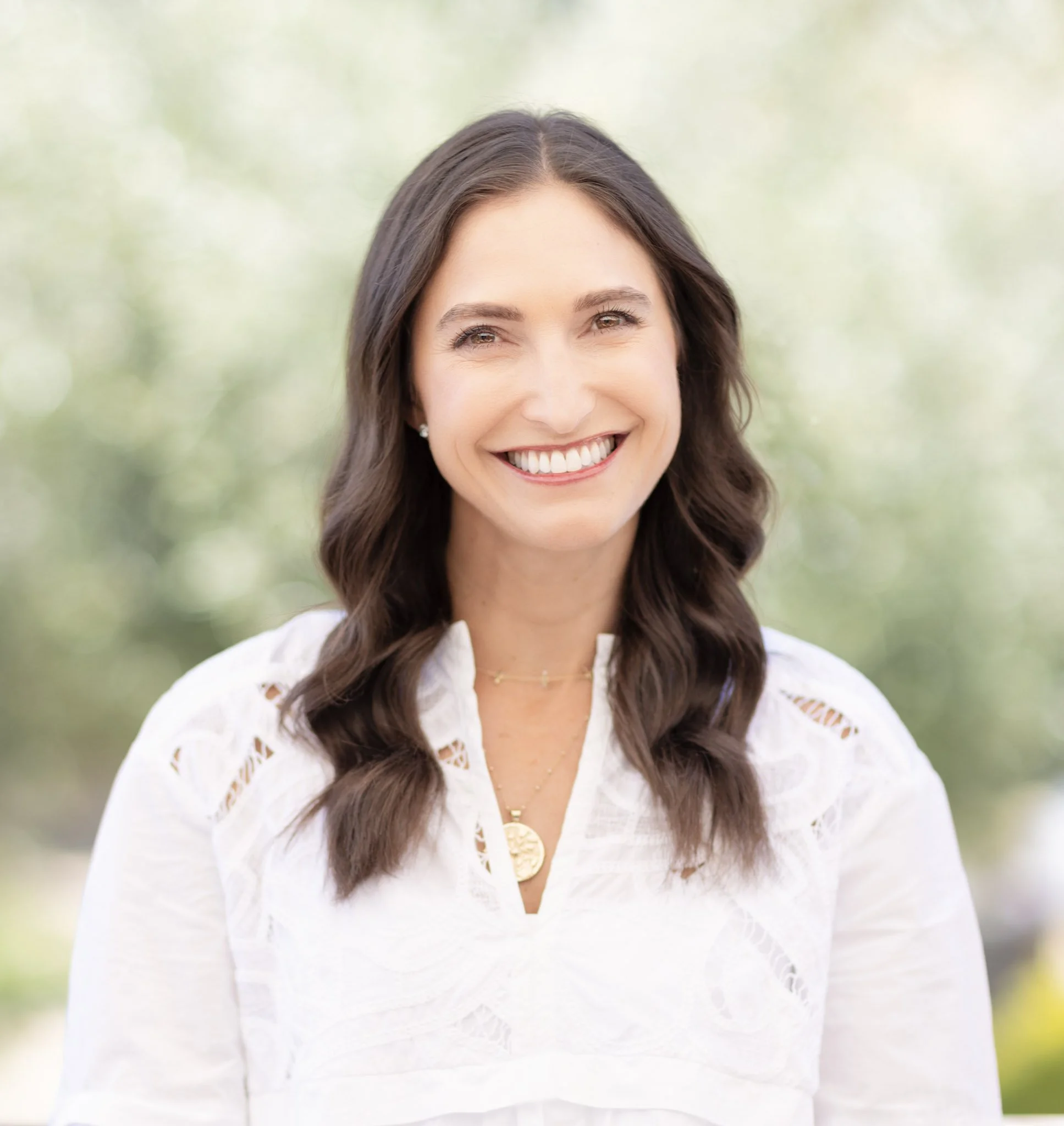 Smiling woman with long dark hair wearing a white blouse and gold jewelry outdoors.