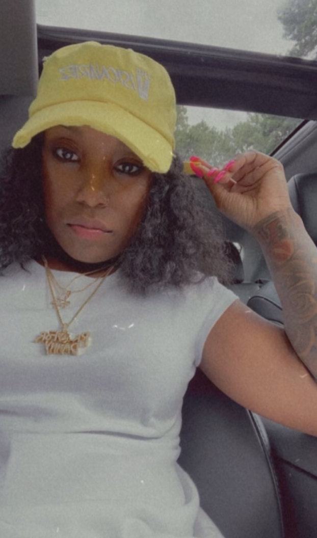 A woman sitting in a car, wearing a yellow cap, white shirt, and gold necklaces, with long curly hair and pink painted nails. The Visionariez'