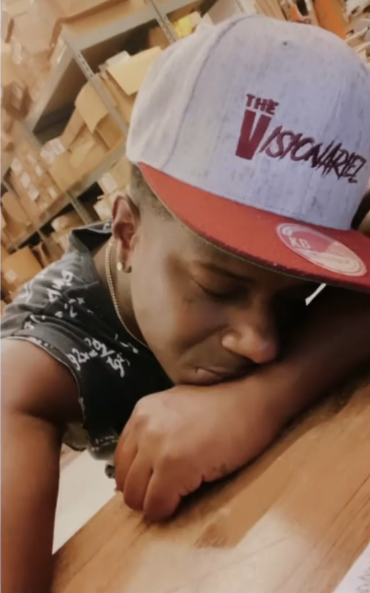 A person resting their head and arm on a wooden surface, wearing a gray and red cap with the words 'The Visionariez,' in a storage room with shelves filled with boxes in the background.