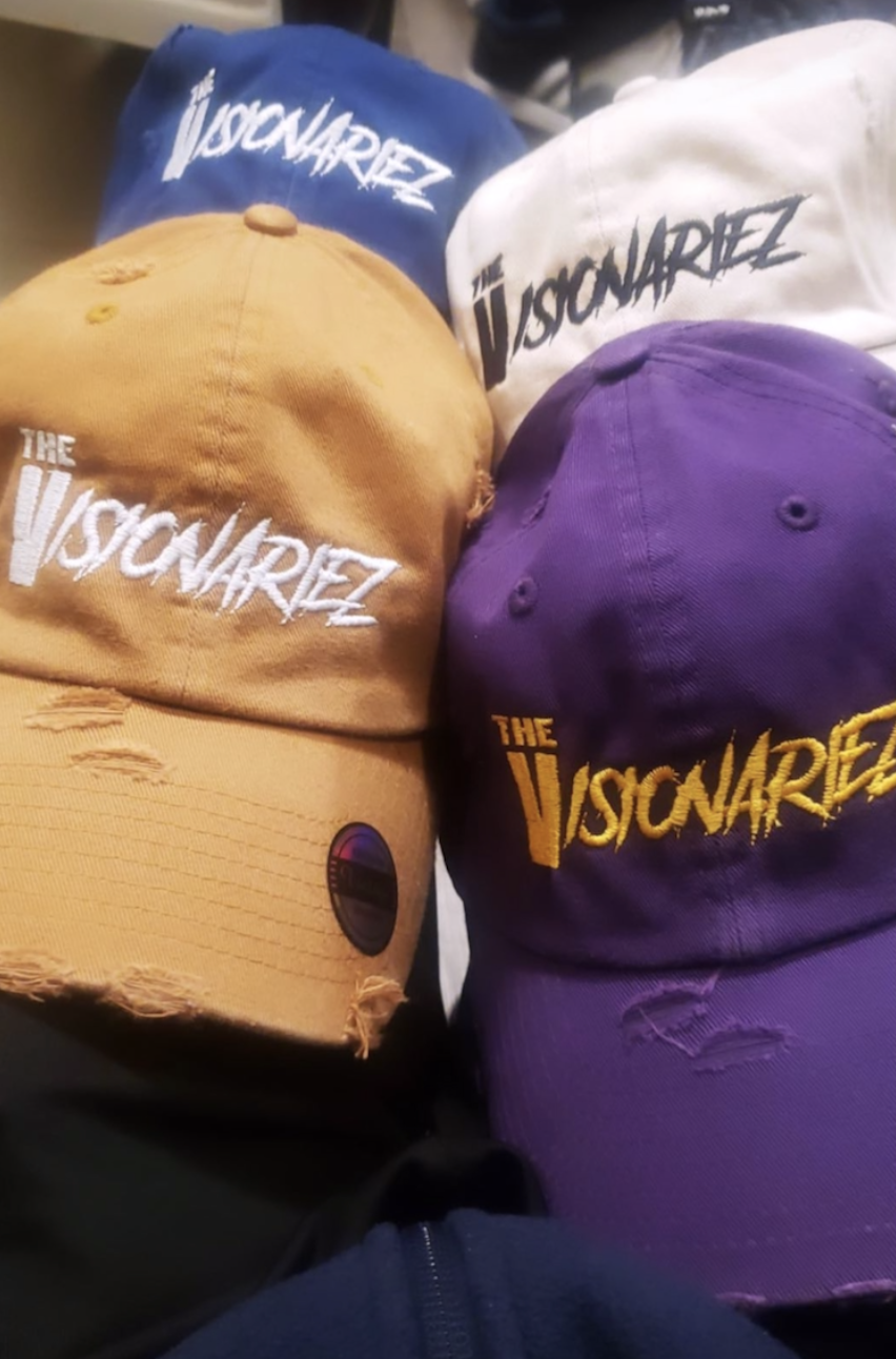 Four baseball caps with the logo and text 'The Visconarez' in different colors: blue, white, orange, and purple, positioned on top of each other.