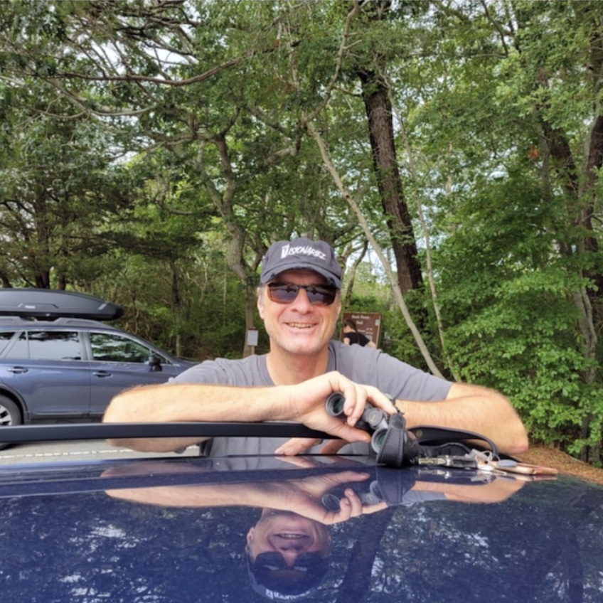 Smiling man with sunglasses and a cap holding binoculars, leaning on a car with a reflected face, in a wooded outdoor area. The Visionariez
