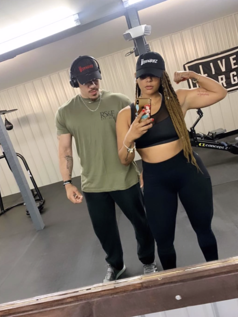 A woman with long braids and a black sports bra flexing her right arm, taking a mirror selfie with a smartphone, standing next to a man in a gym with workout equipment and a sign on the wall that says 'LIVE'. Both are wearing black caps and athletic