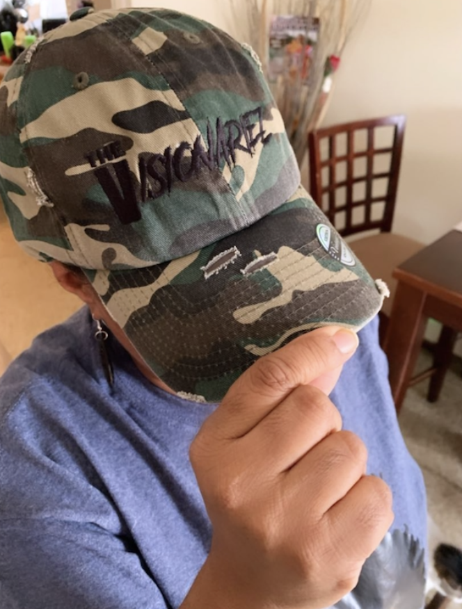 Person wearing a camouflage baseball cap with black embroidered text that reads 'The Visionariez'', holding the brim of the cap.