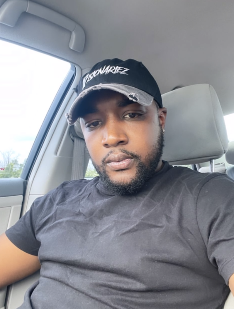 A man with a beard wearing a black T-shirt and a black baseball cap The Visionariez' sitting in the passenger seat of a car