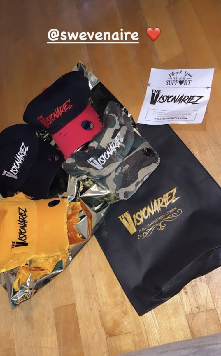 Multiple caps and a black gift bag with "The Visionaryz" logo and slogan, placed on a wooden floor alongside a piece of paper with a message. The caps are black, red, and camouflage, all with the same logo.