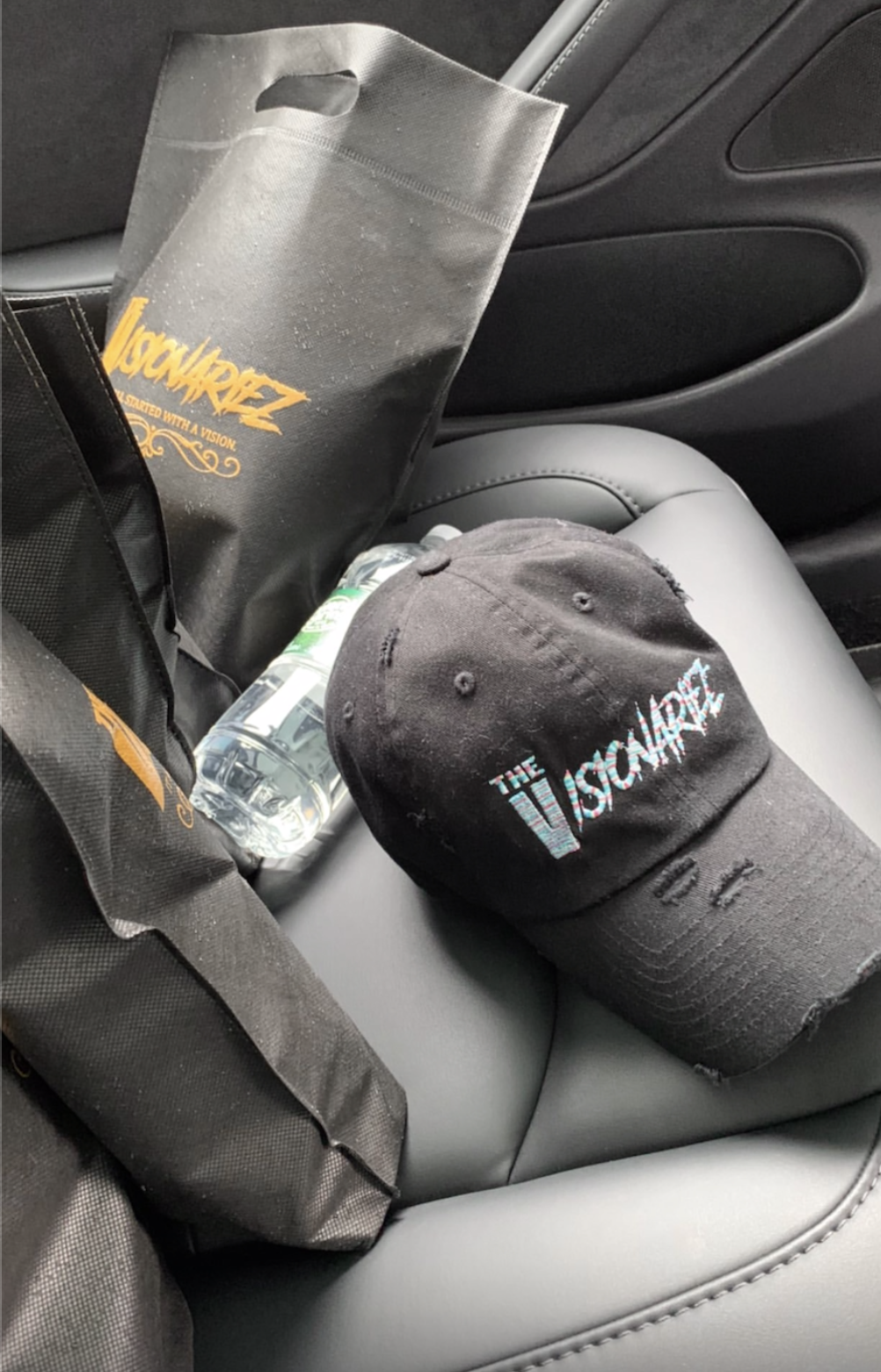 A black cap with colorful embroidered text reading 'The Visionariez' resting on a car seat. A water bottle and a black tote bag attached to a black backpack are also on the seat.