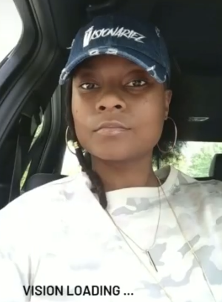 Woman with hoop earrings wearing a white camouflage shirt and a navy blue cap with the word 'The Visionariez'' on it, sitting in a vehicle.