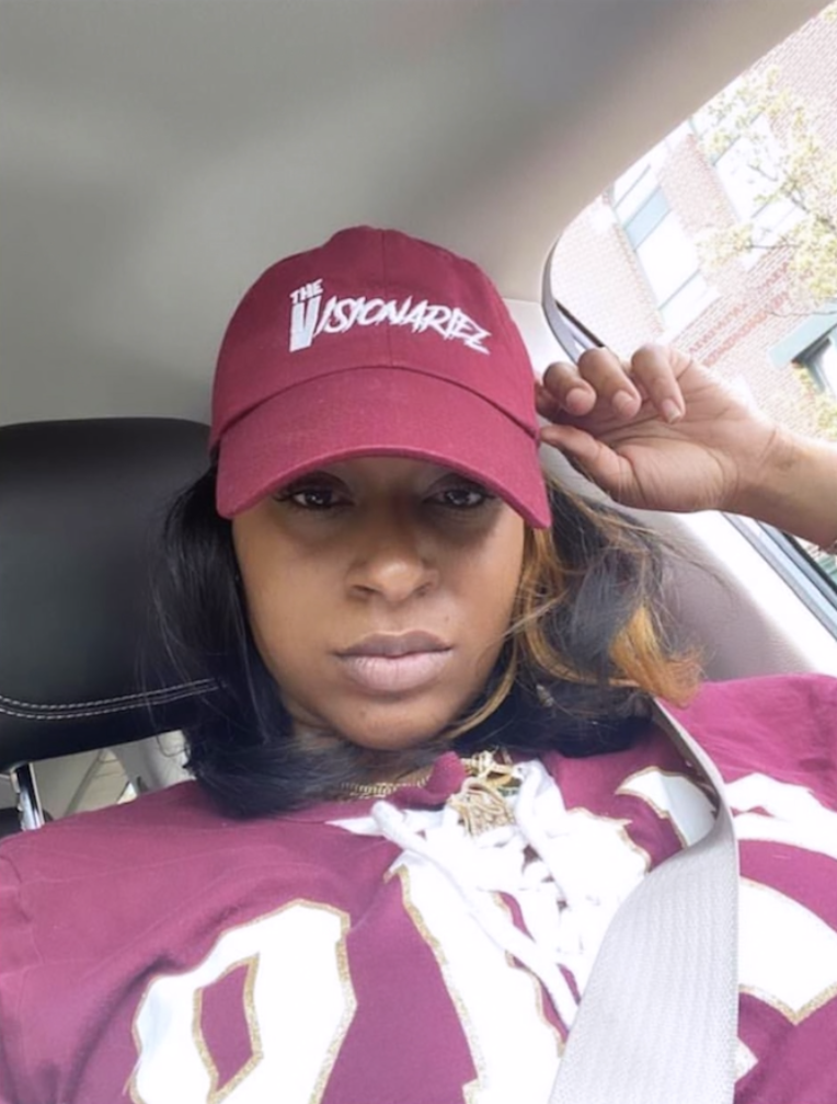 Woman wearing a burgundy cap and sports jersey, sitting in a car, with her left hand adjusting the cap The Visionariez'.